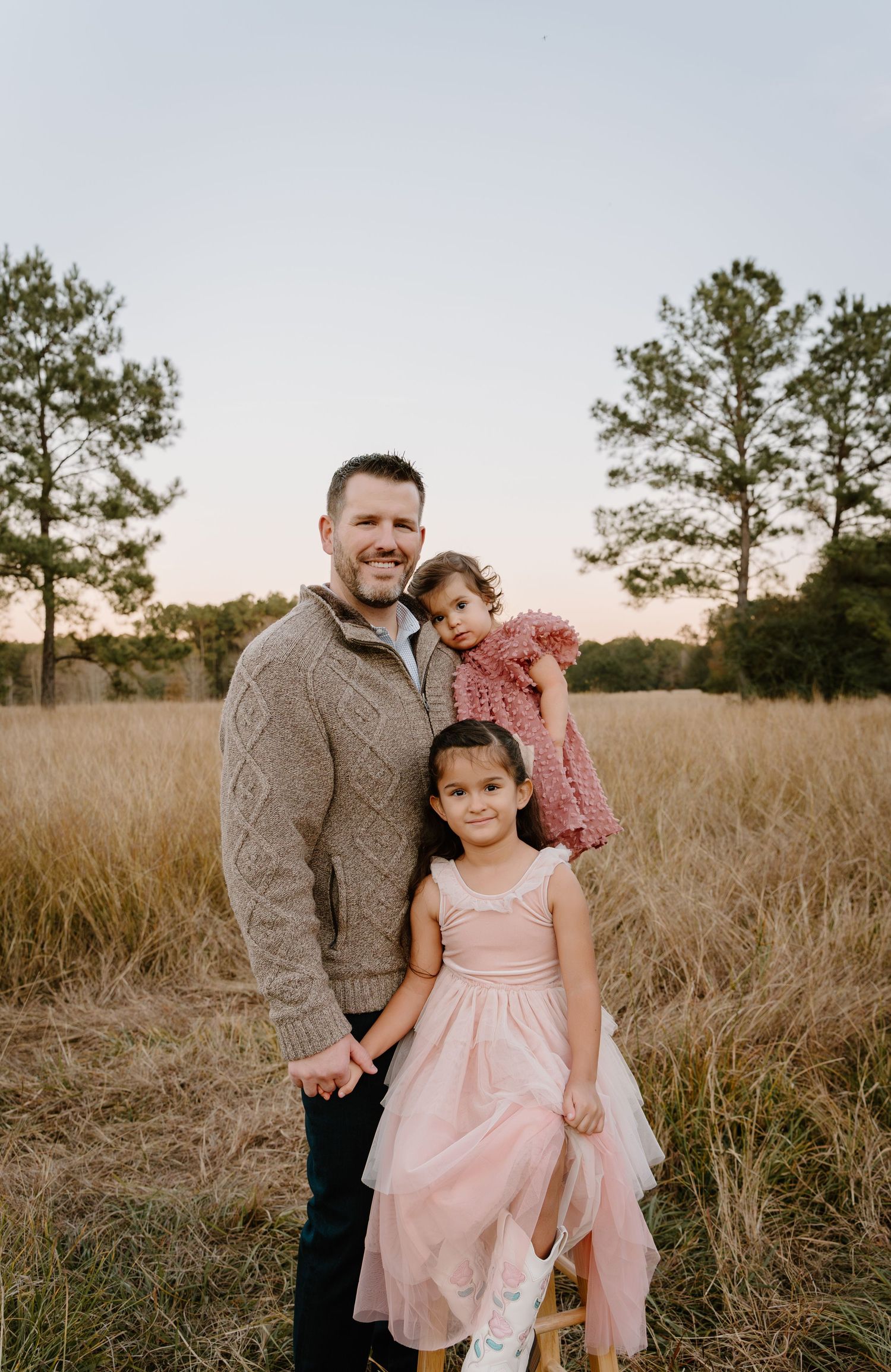 A father smiles with two young daughters in pink dresses in a golden field at sunset.