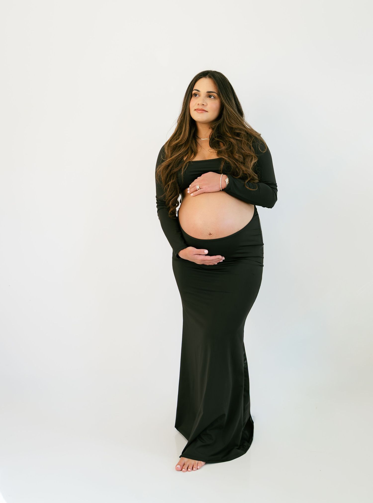 Pregnant woman in black off-shoulder maxi dress cradling baby bump, posing barefoot against white studio background.