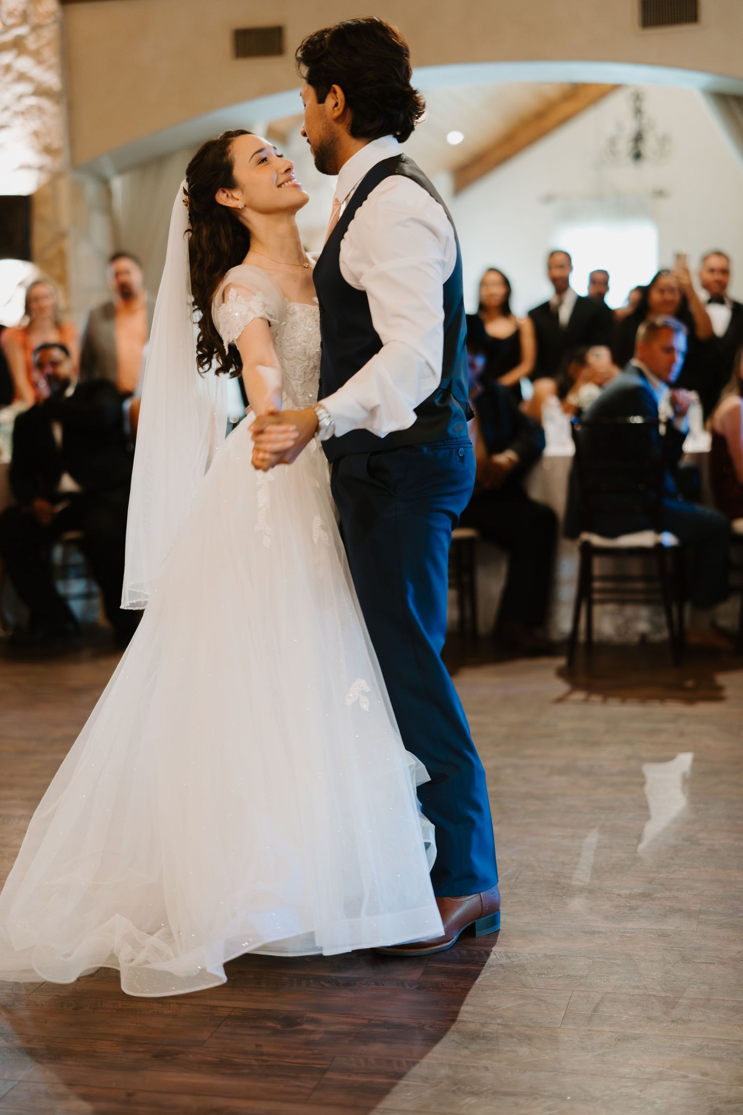 Bride and groom share their first dance as newlyweds while guests watch in a beautifully lit reception venue.
