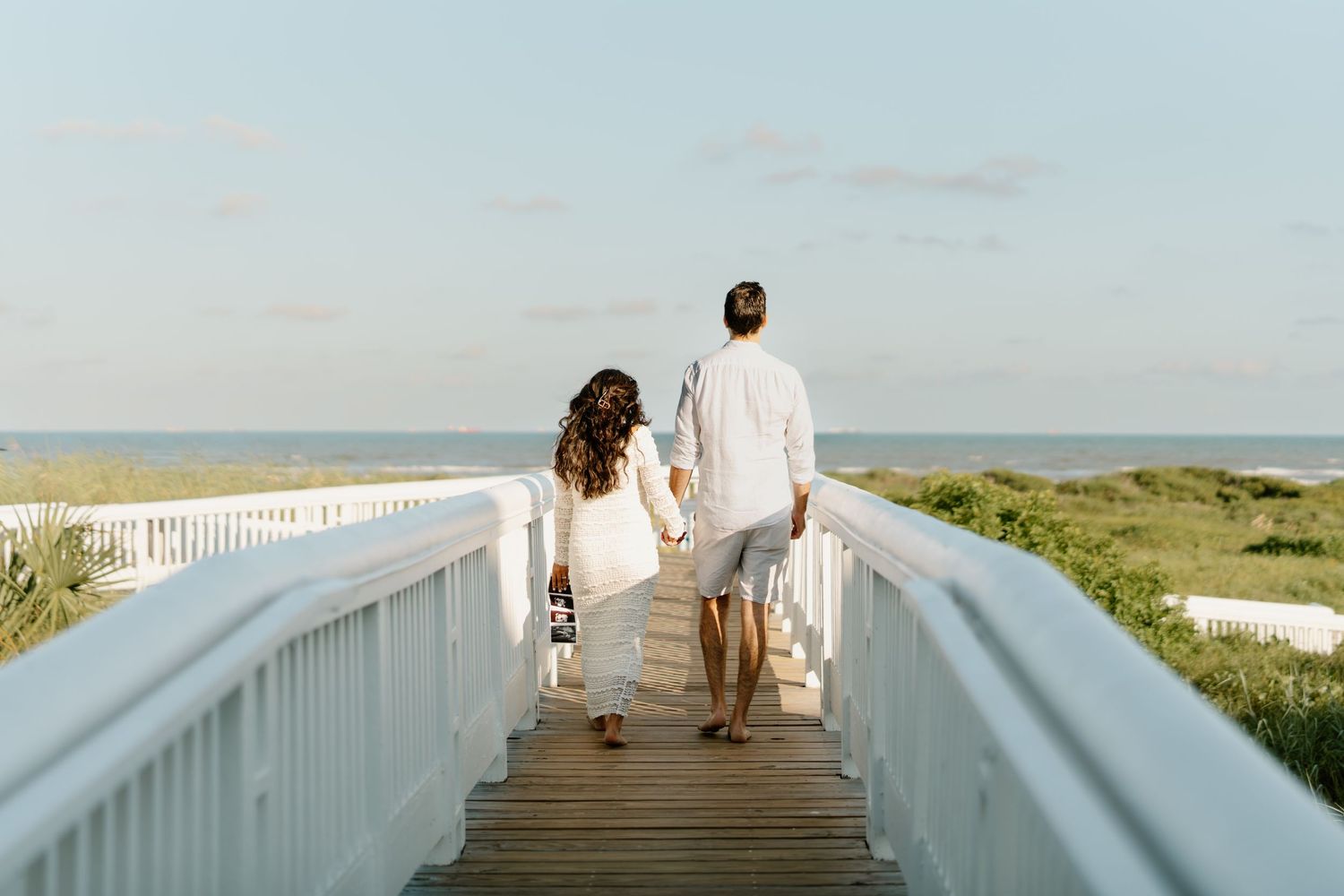 Couple holding hands walking on a wooden boardwalk toward a serene beach at golden hour.