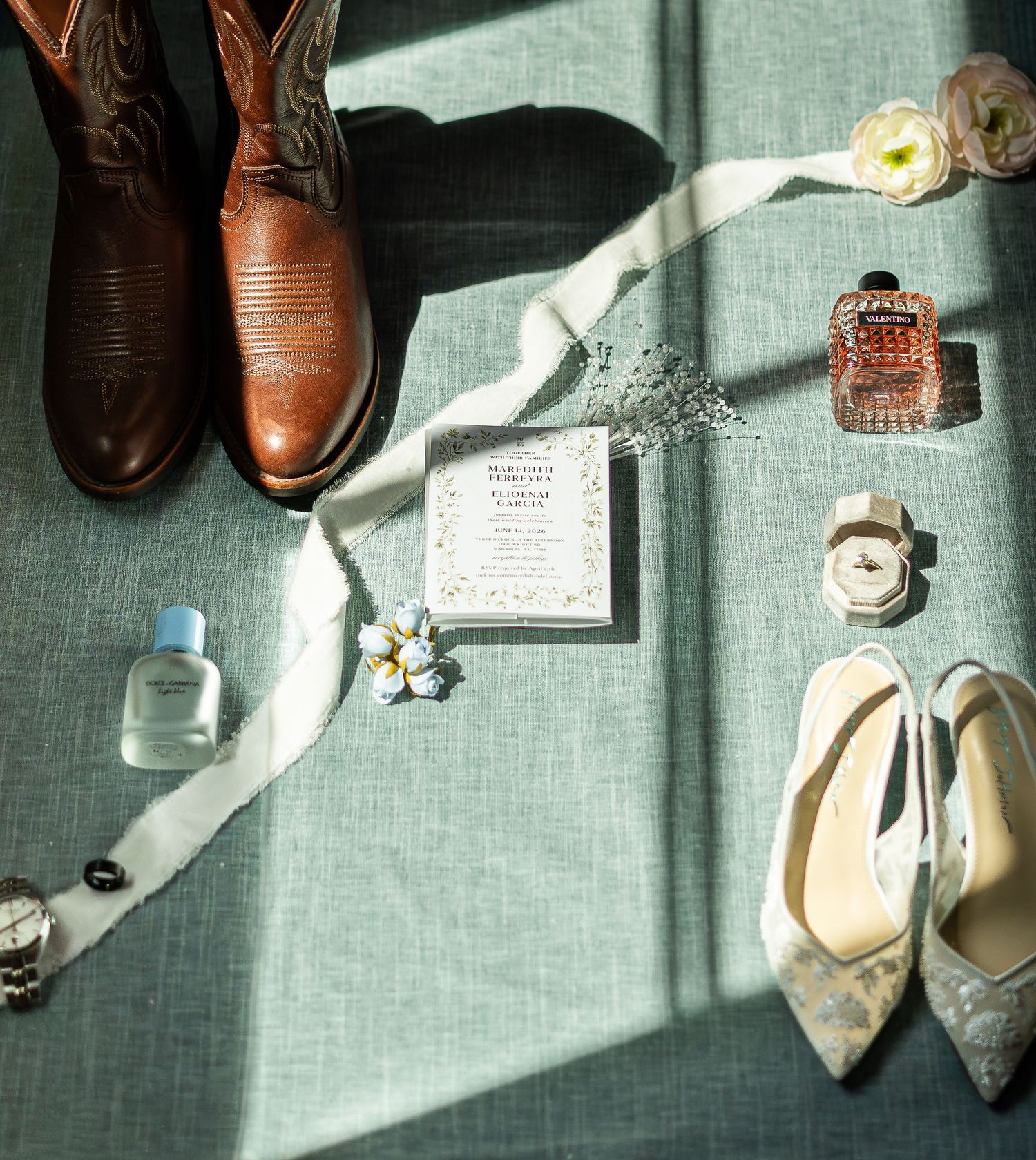 Flat lay of wedding accessories including cowboy boots, heels, invitation, perfume, and floral details on sage linen.