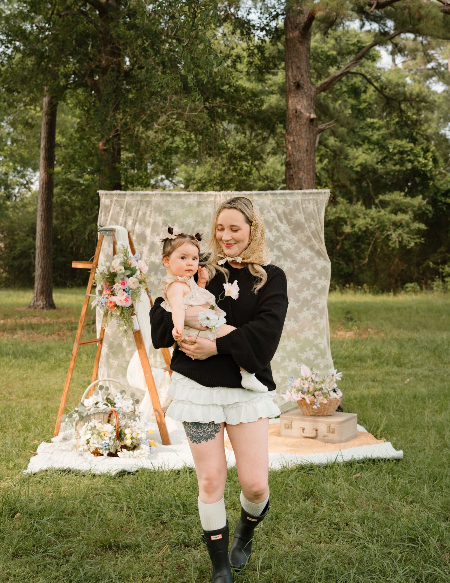 A smiling woman holds a baby in a boho outdoor photo session with floral decor and lace backdrop.