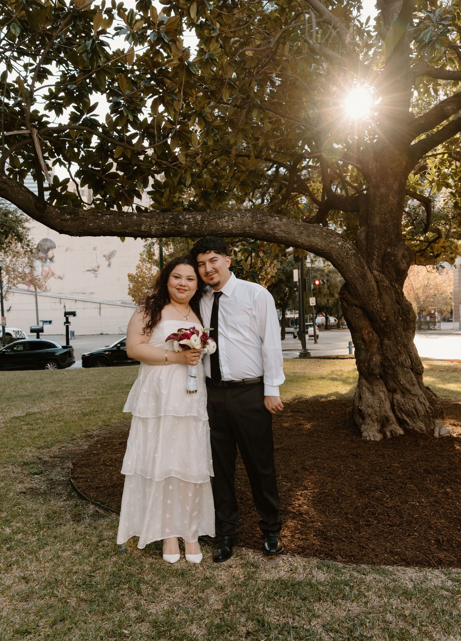 Couple poses under a sunlit magnolia tree, bride in white tiered dress holding red bouquet, groom in black suit.