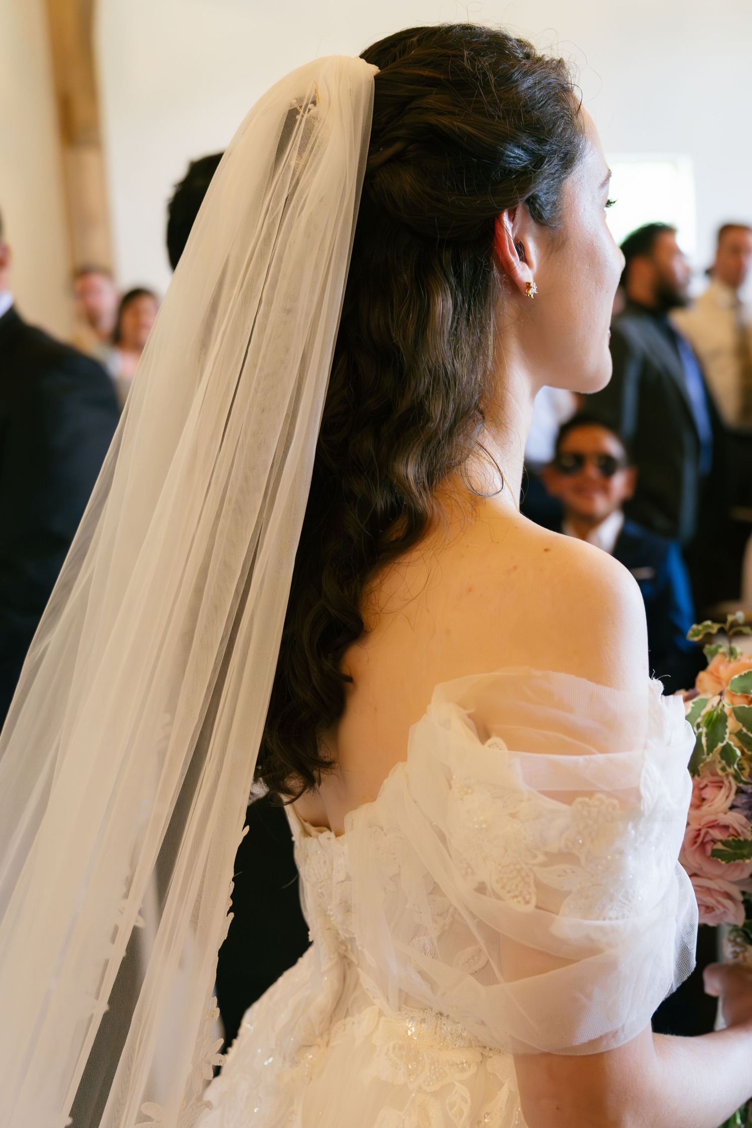 Bride with long curly hair and white veil in off-shoulder wedding gown holding bouquet at ceremony.