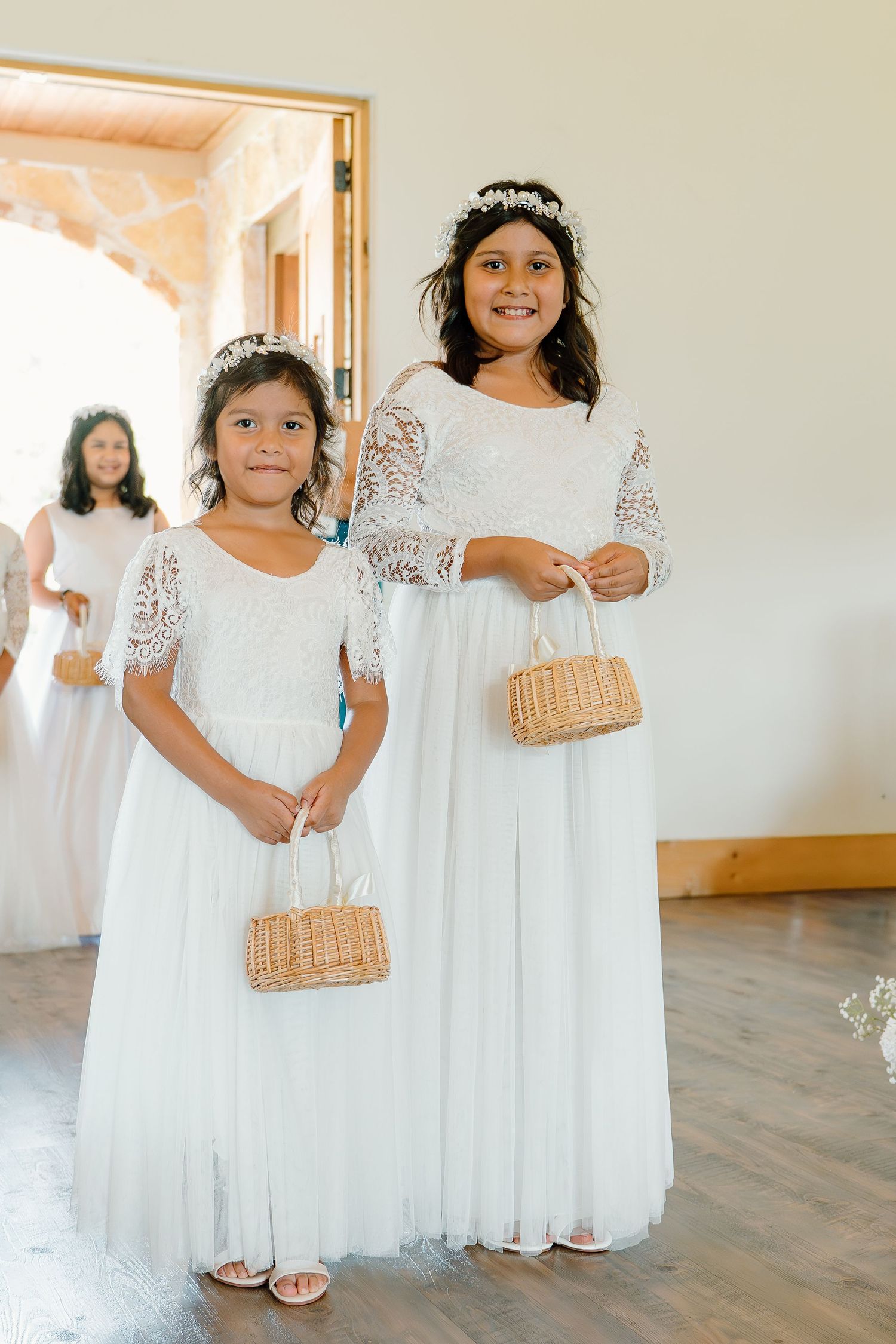 Two flower girls in white lace dresses holding wicker baskets at a wedding ceremony.