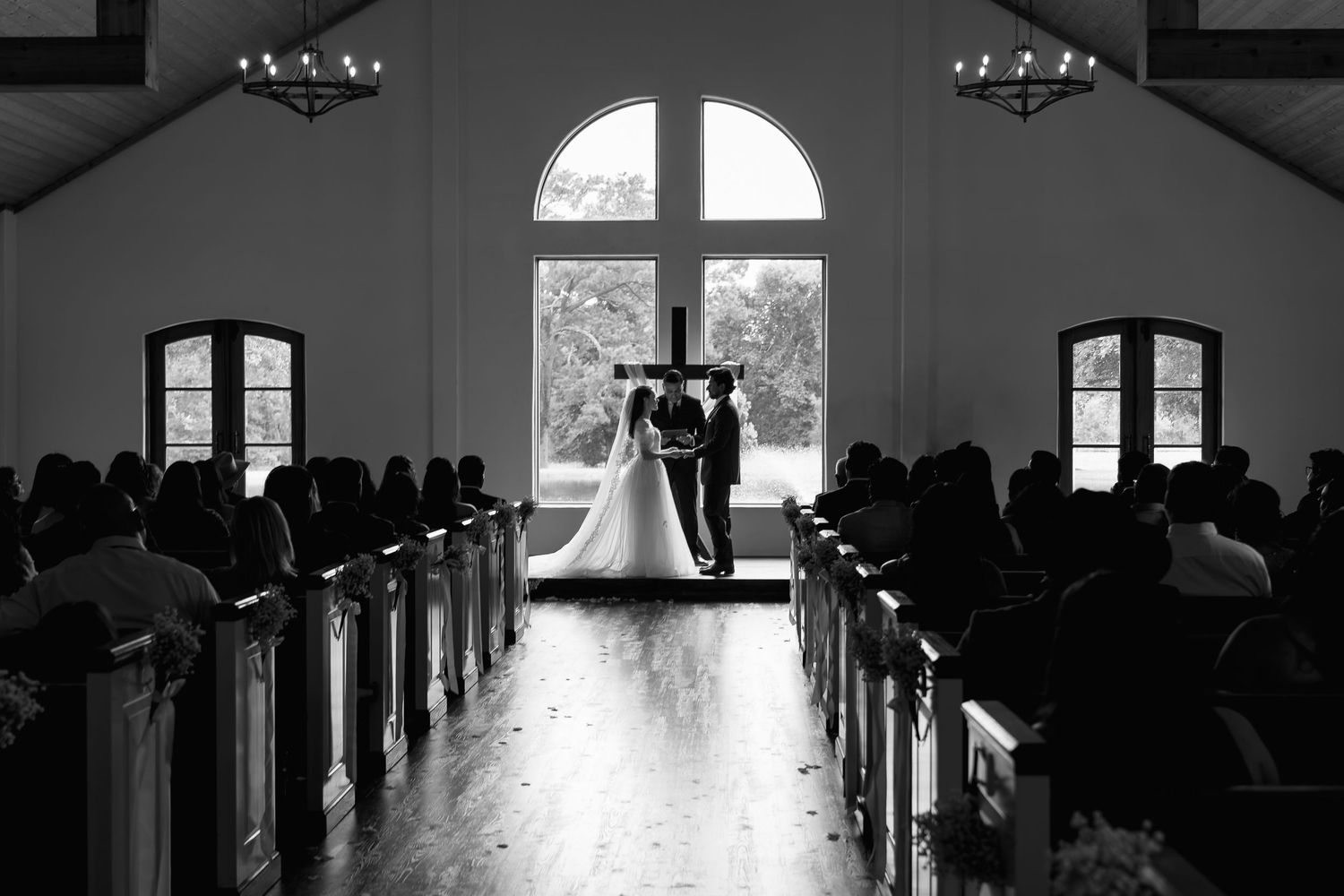 Black and white photo of a bride and groom at the altar of a chapel with large arched windows and wooden pews.