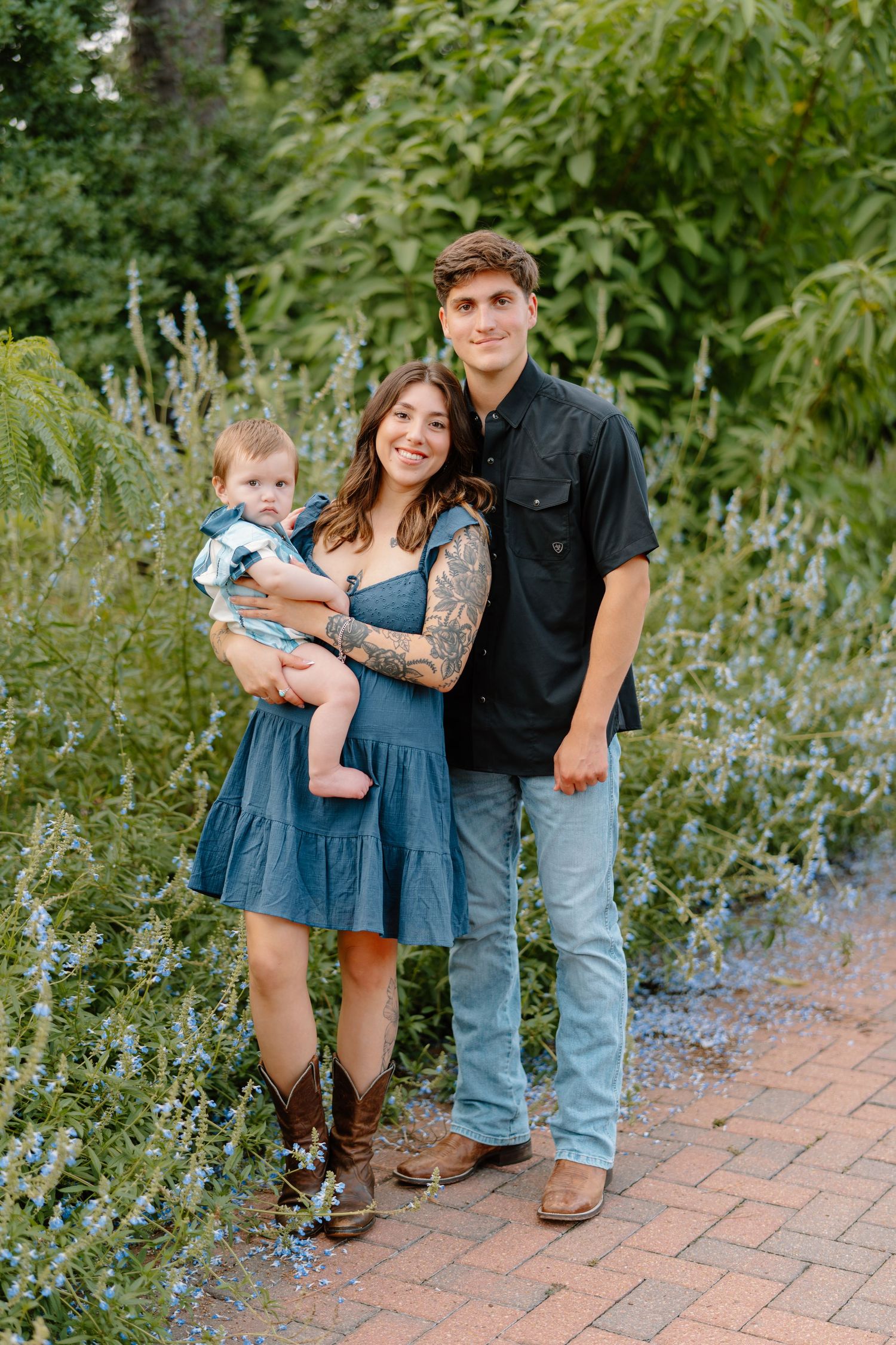 Young family of three poses outdoors, woman in blue dress holding baby, man in black shirt, surrounded by lush greenery.