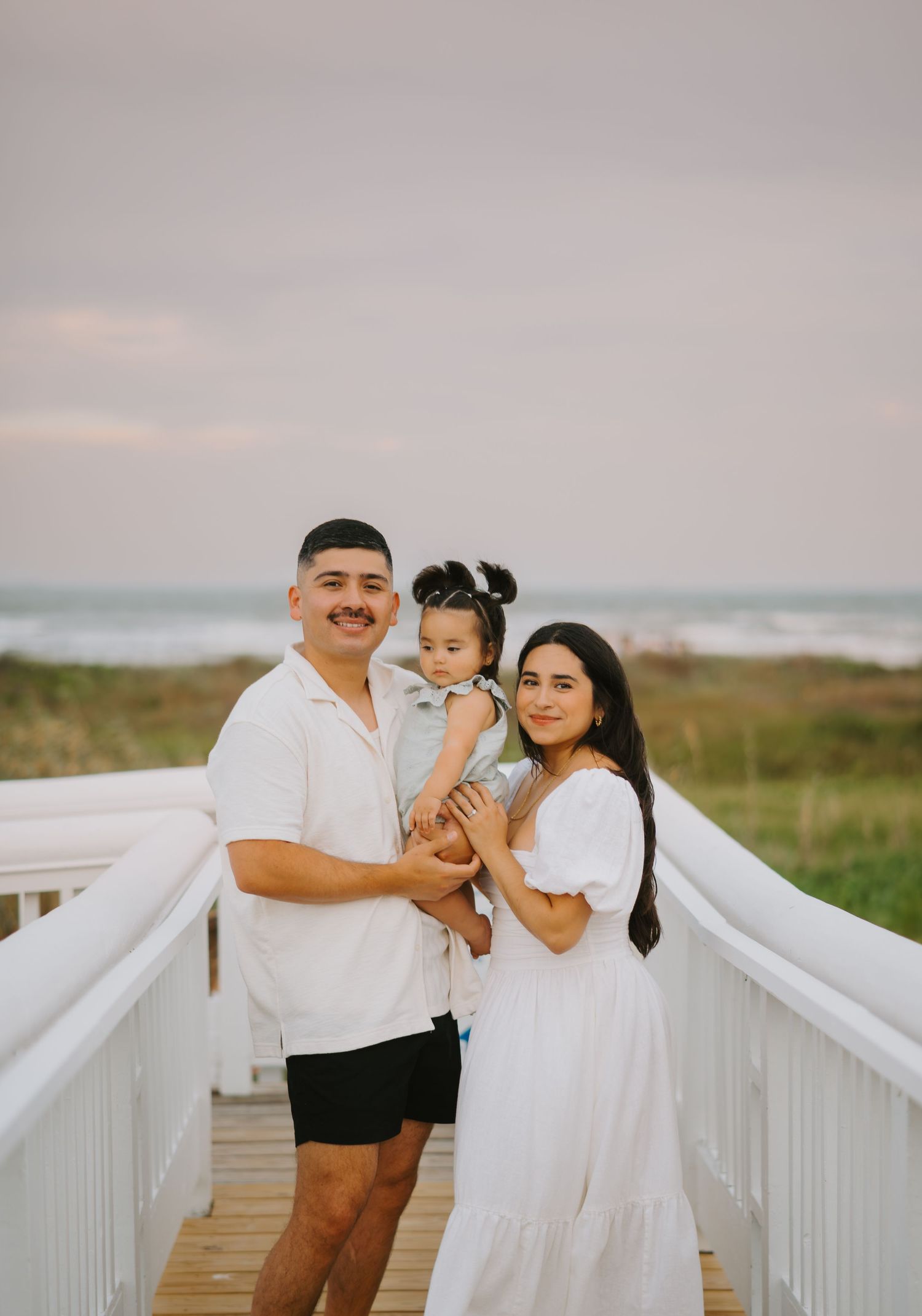 A young family of three poses on a white wooden beach boardwalk, with parents holding their toddler against an ocean backdrop.