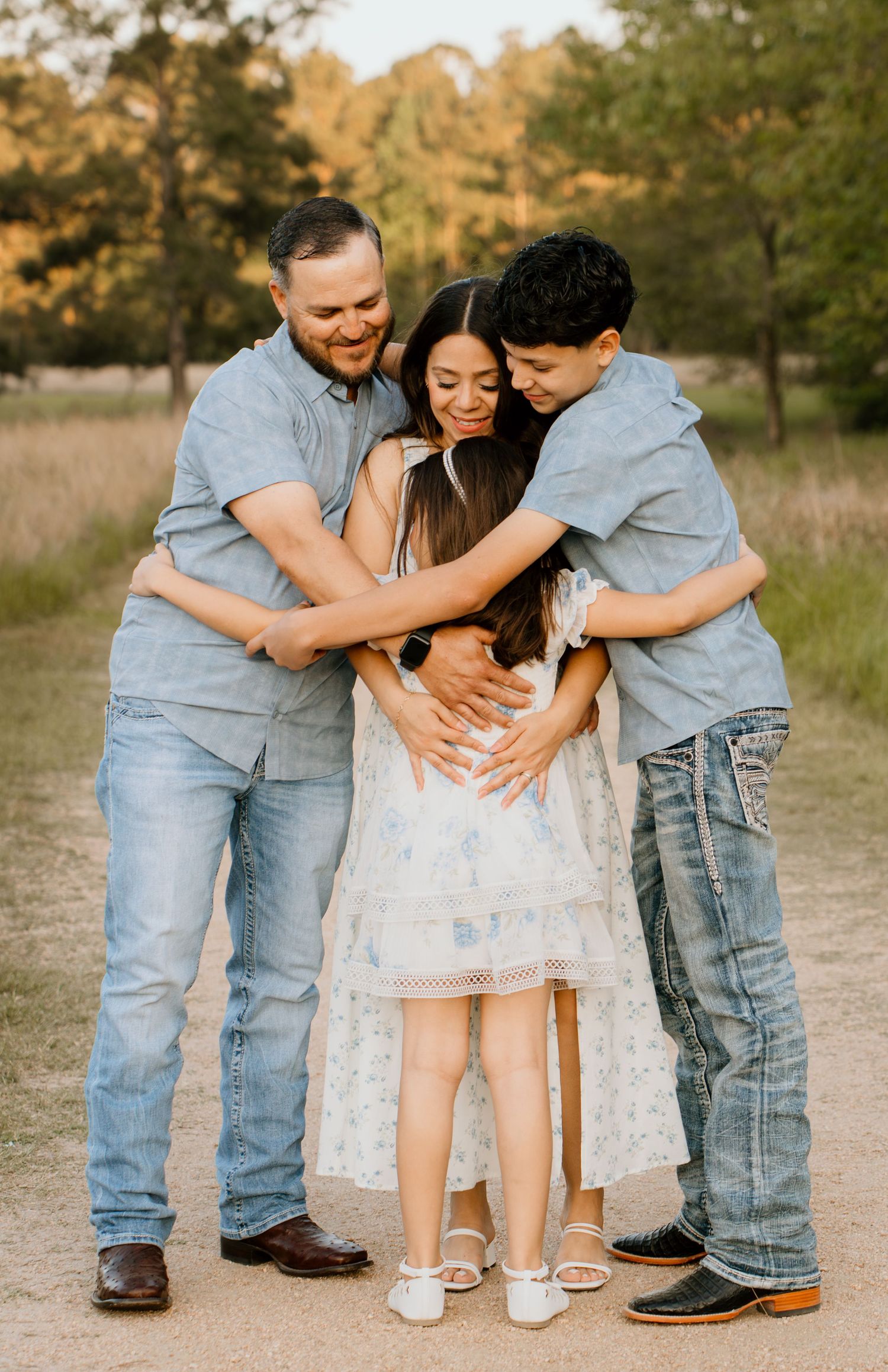 Family of four sharing a warm group hug on a dirt path, dressed in blue and white, surrounded by golden trees.