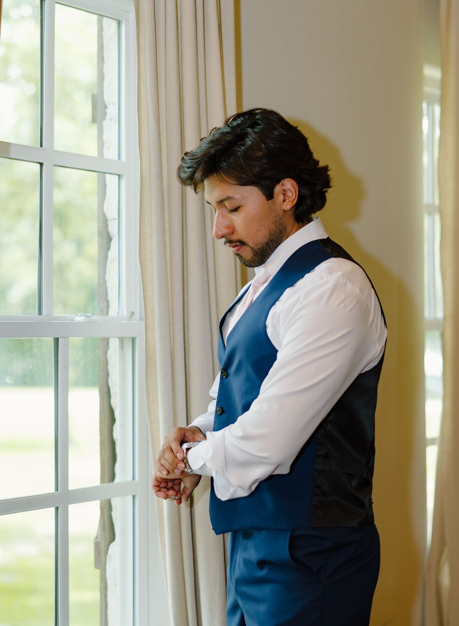Groom in blue vest and white shirt adjusting cufflinks by a bright window before wedding ceremony.