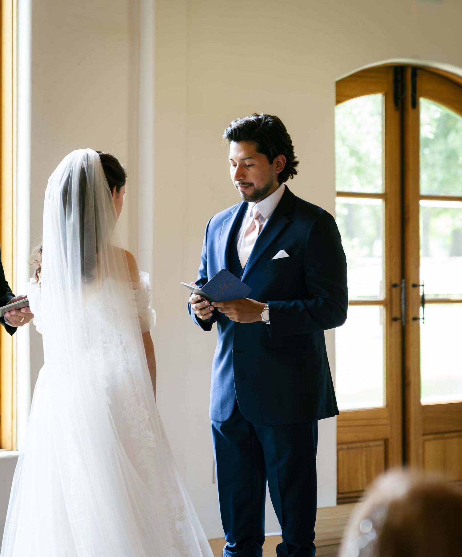 Groom in navy blue suit reads wedding vows to bride in white lace gown during elegant indoor ceremony.