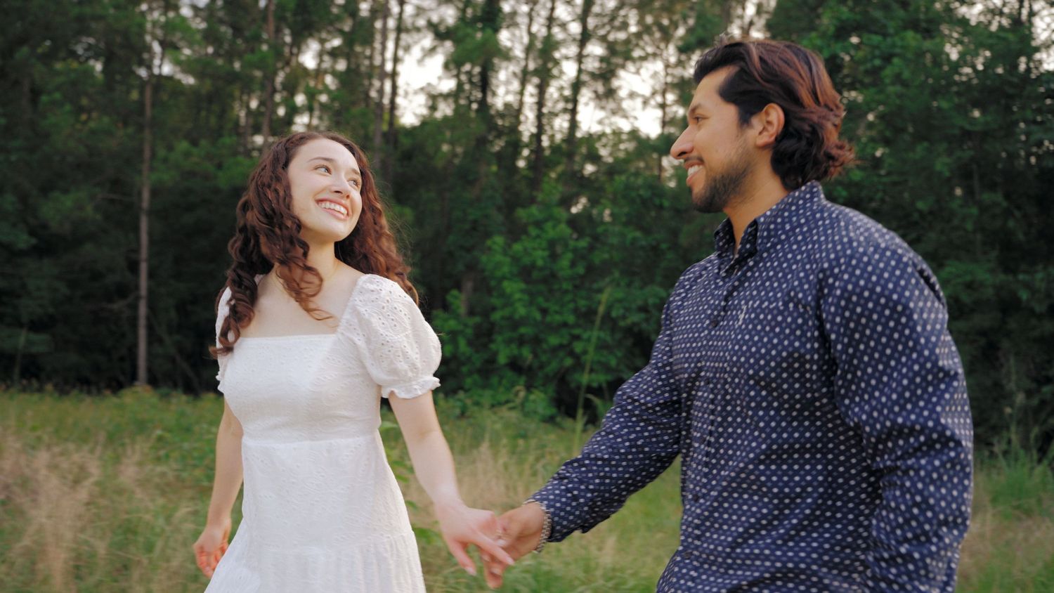 A smiling couple holds hands in a lush green meadow, woman in white dress, man in blue shirt.