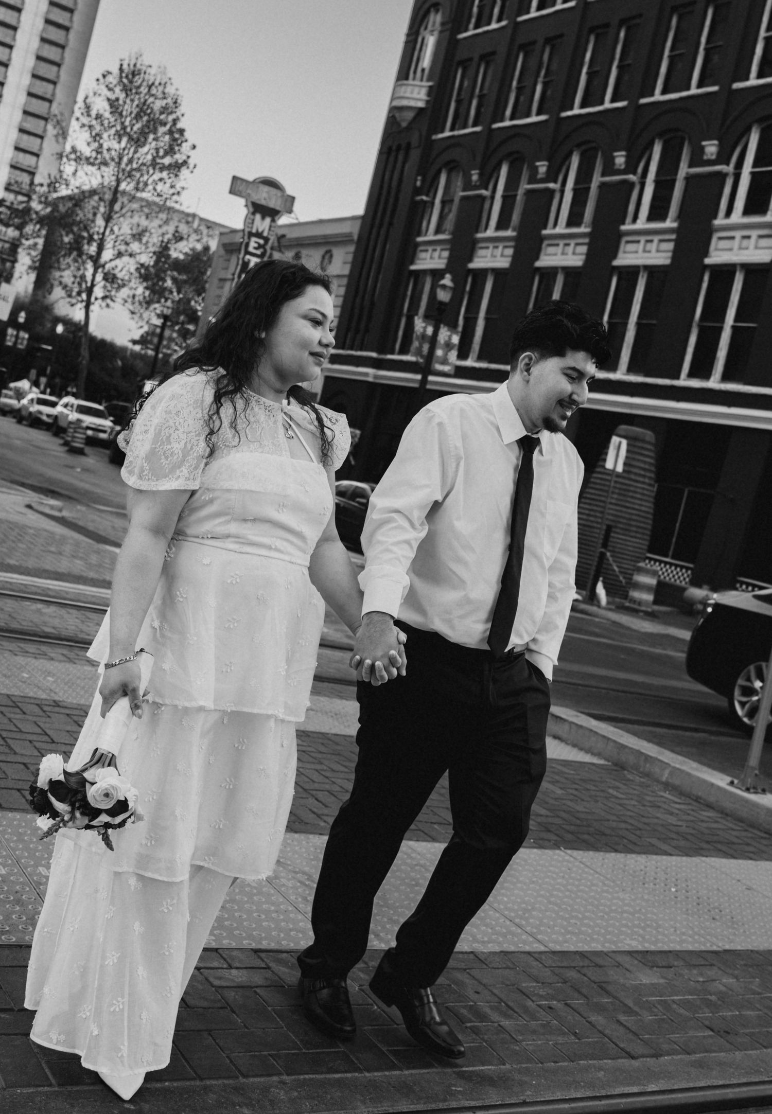 Black and white photo of bride in white dress and groom in white shirt crossing city street holding hands.