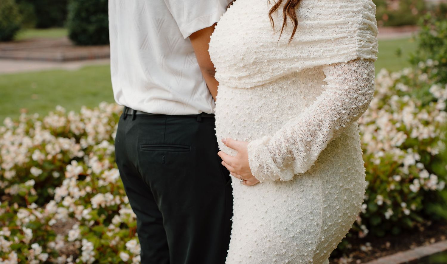 Pregnant couple embracing outdoors in front of white flowers, woman wearing elegant white textured maternity dress.
