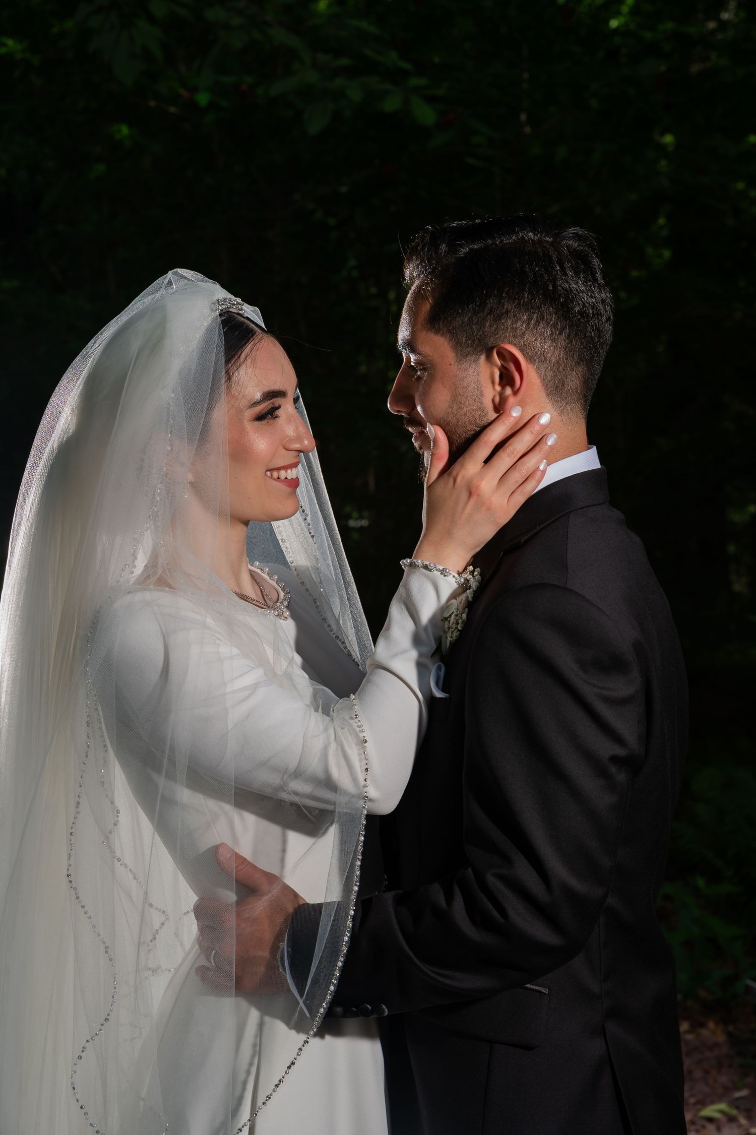 A smiling bride in white gown and veil tenderly touches groom's face as they share a romantic moment outdoors.