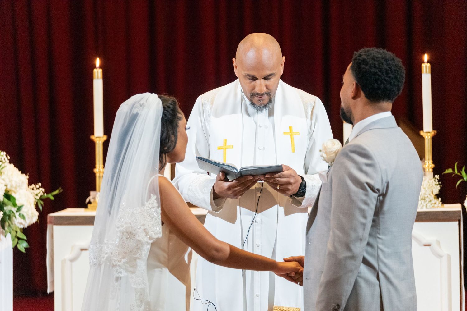 A bride and groom exchange vows before a officiant at a church wedding altar with candles and flowers.