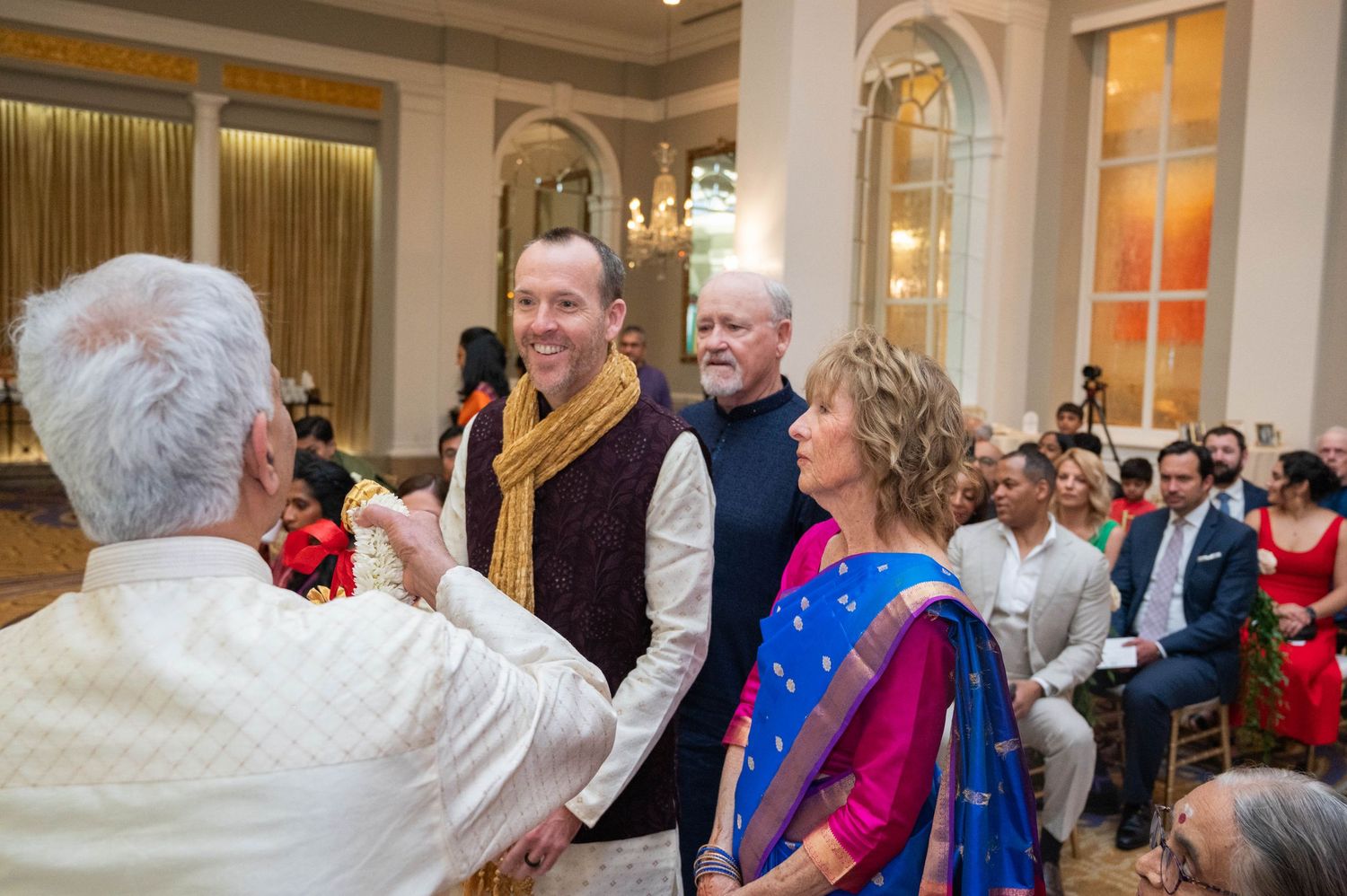Guests in formal and traditional Indian attire smile and greet each other at an elegant indoor wedding reception.