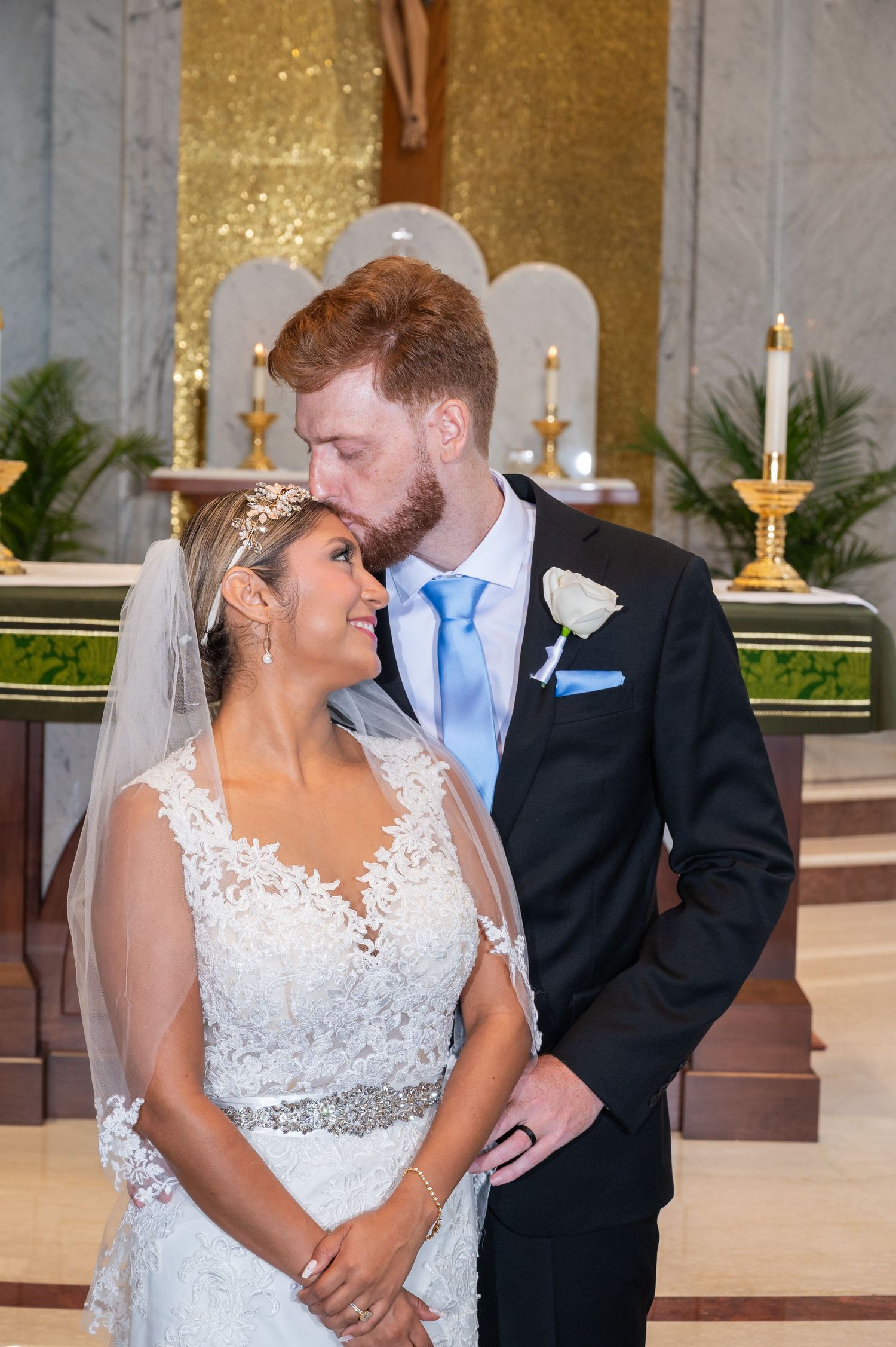 A groom in a dark suit kisses his bride on the forehead at a church altar, as she smiles in a lace wedding gown.