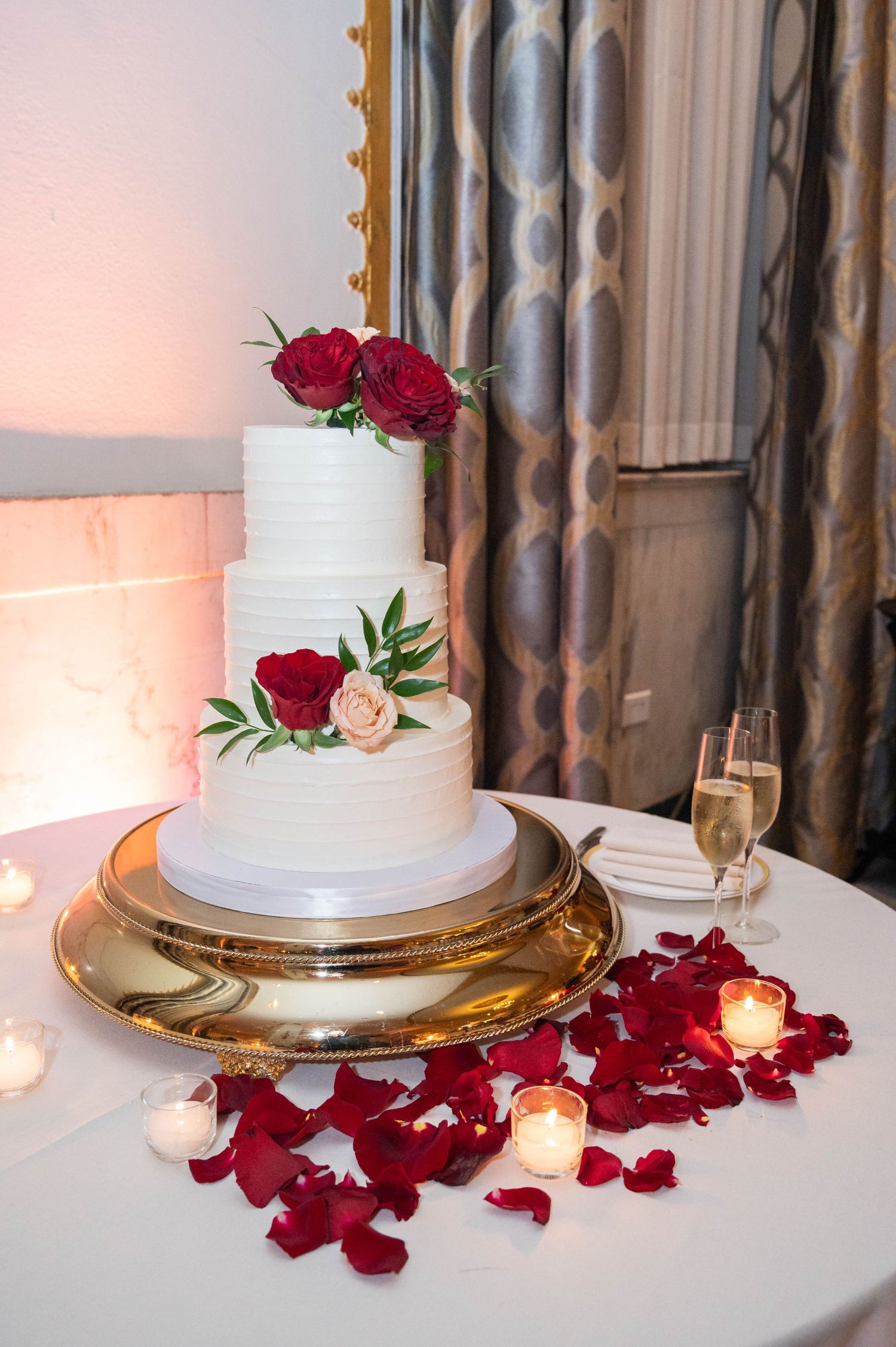 Three-tier white wedding cake with red roses on gold stand surrounded by rose petals.