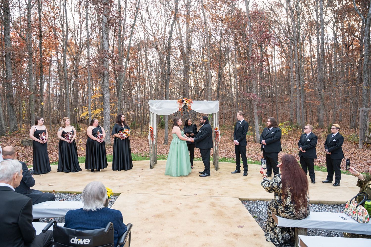 Outdoor fall wedding ceremony with bridal party in black dresses on a patio surrounded by autumn trees.