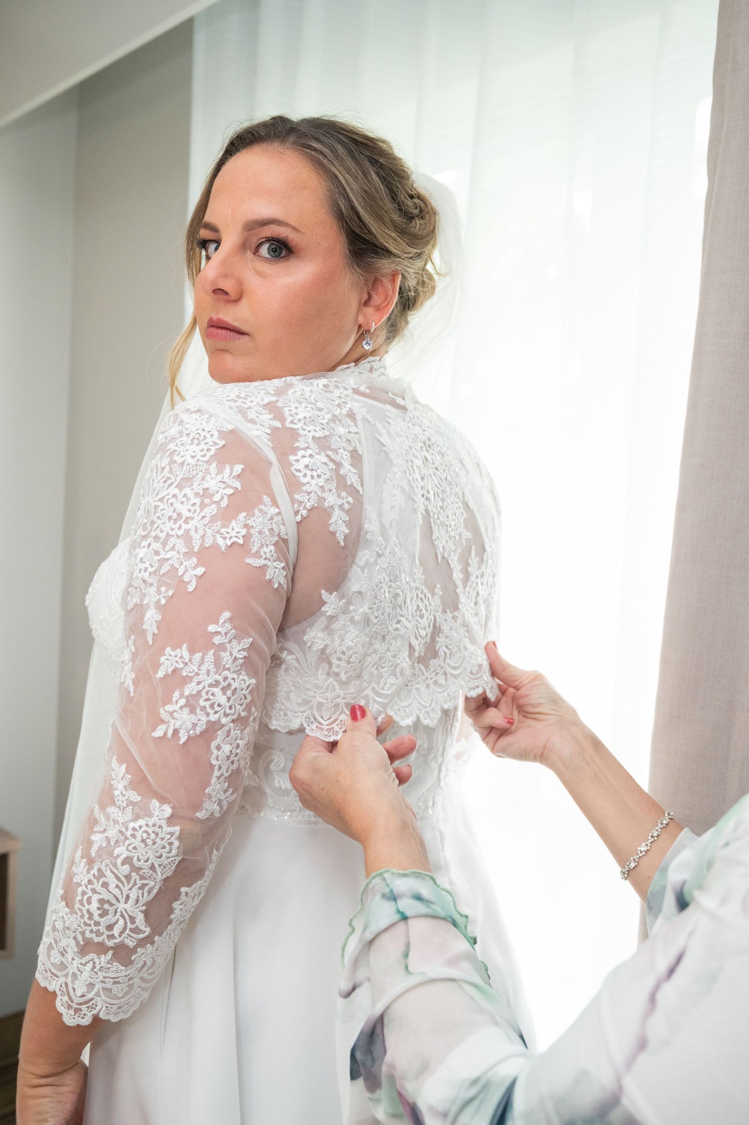 Bride in elegant lace wedding gown being buttoned up by an assistant near a sunlit window.