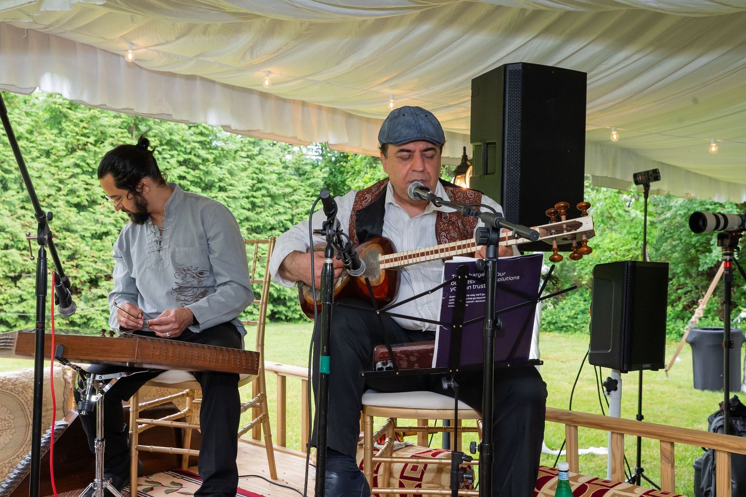 Two musicians perform under a tent, one playing keyboard and another playing a stringed instrument at an outdoor event.
