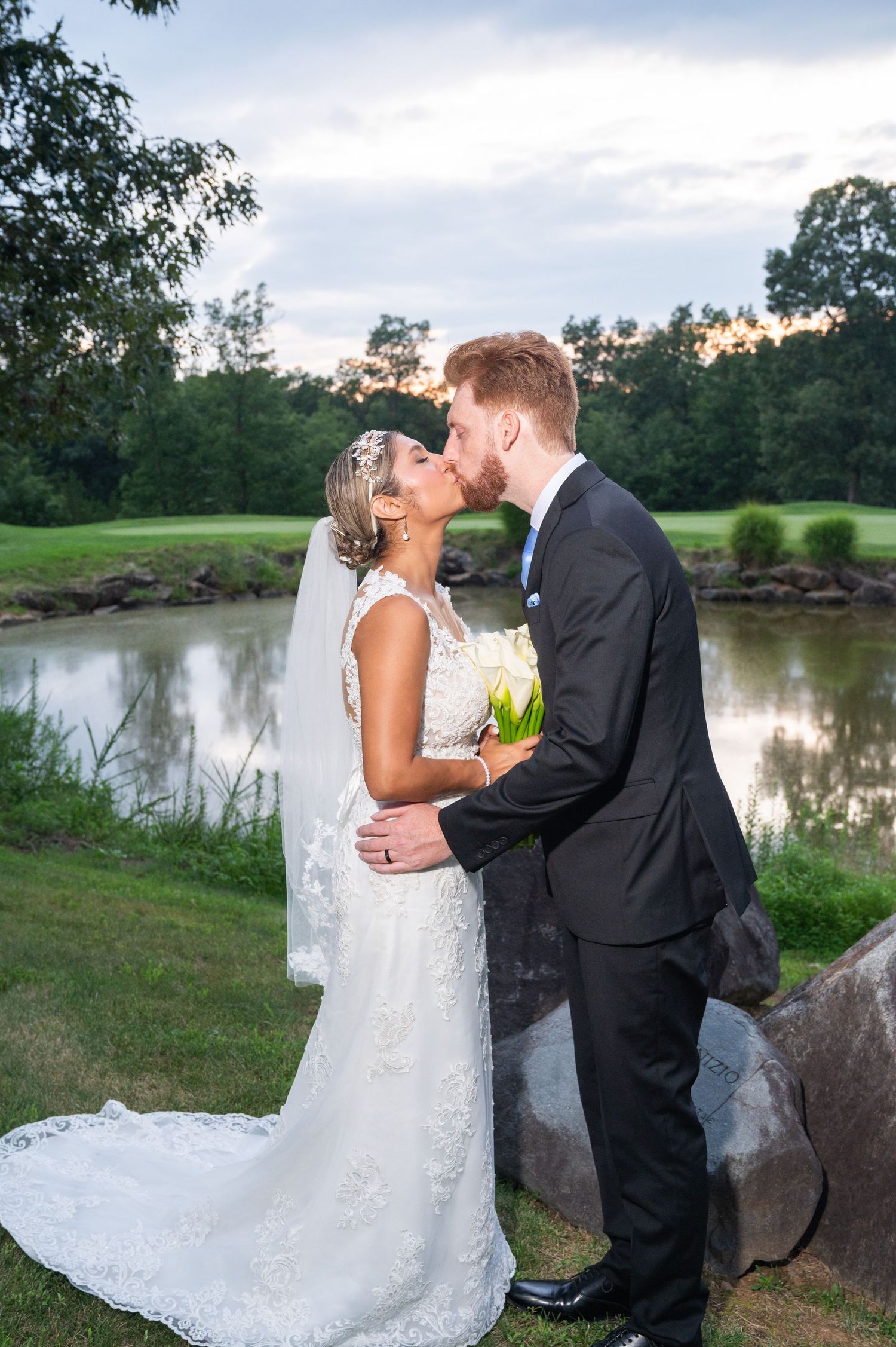Newlyweds kiss by a serene pond at dusk, bride in lace gown with veil, groom in black suit.