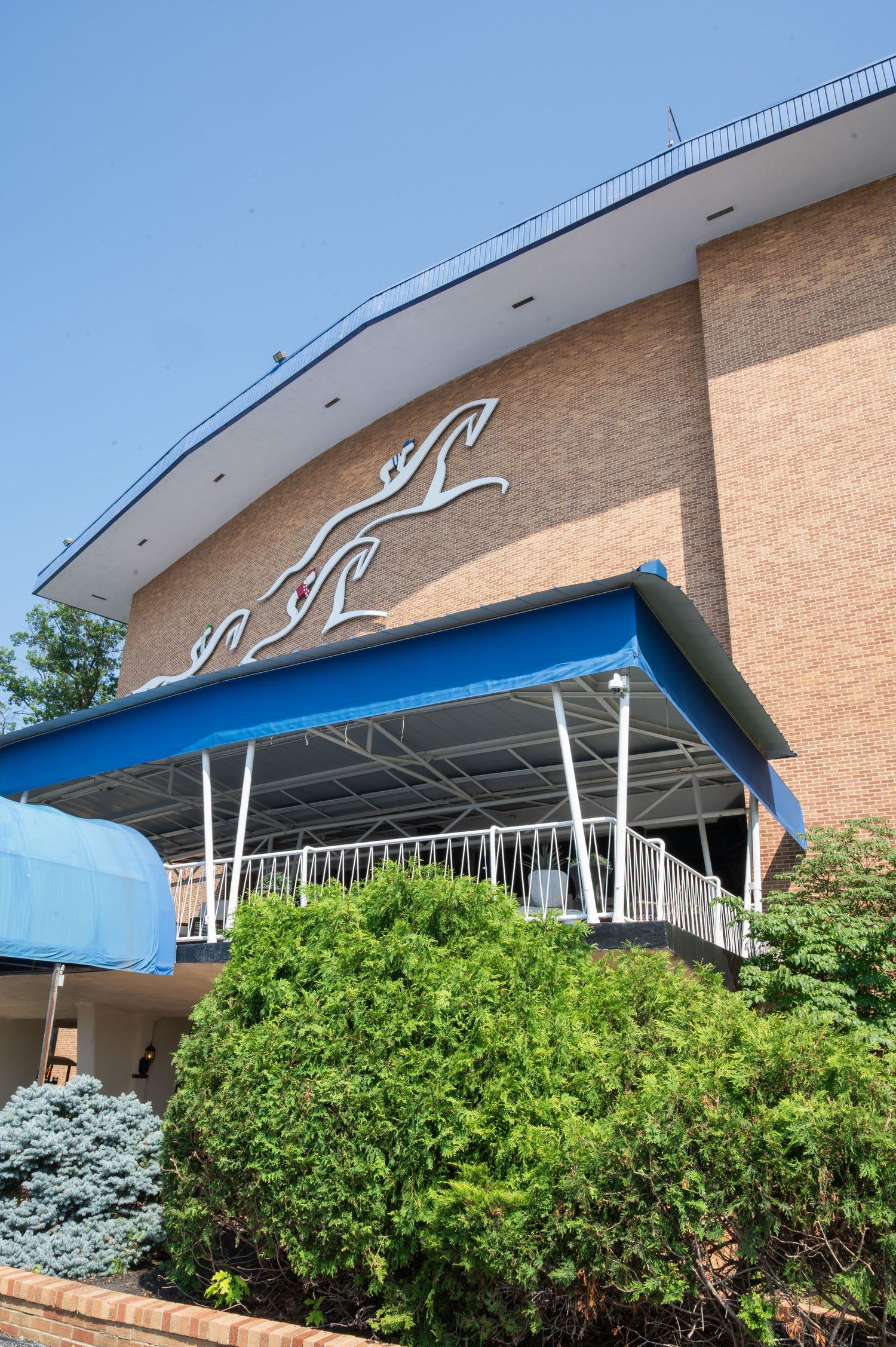 Brick building exterior with blue awning canopy, decorative horse logo, and lush green shrubs under clear blue sky.