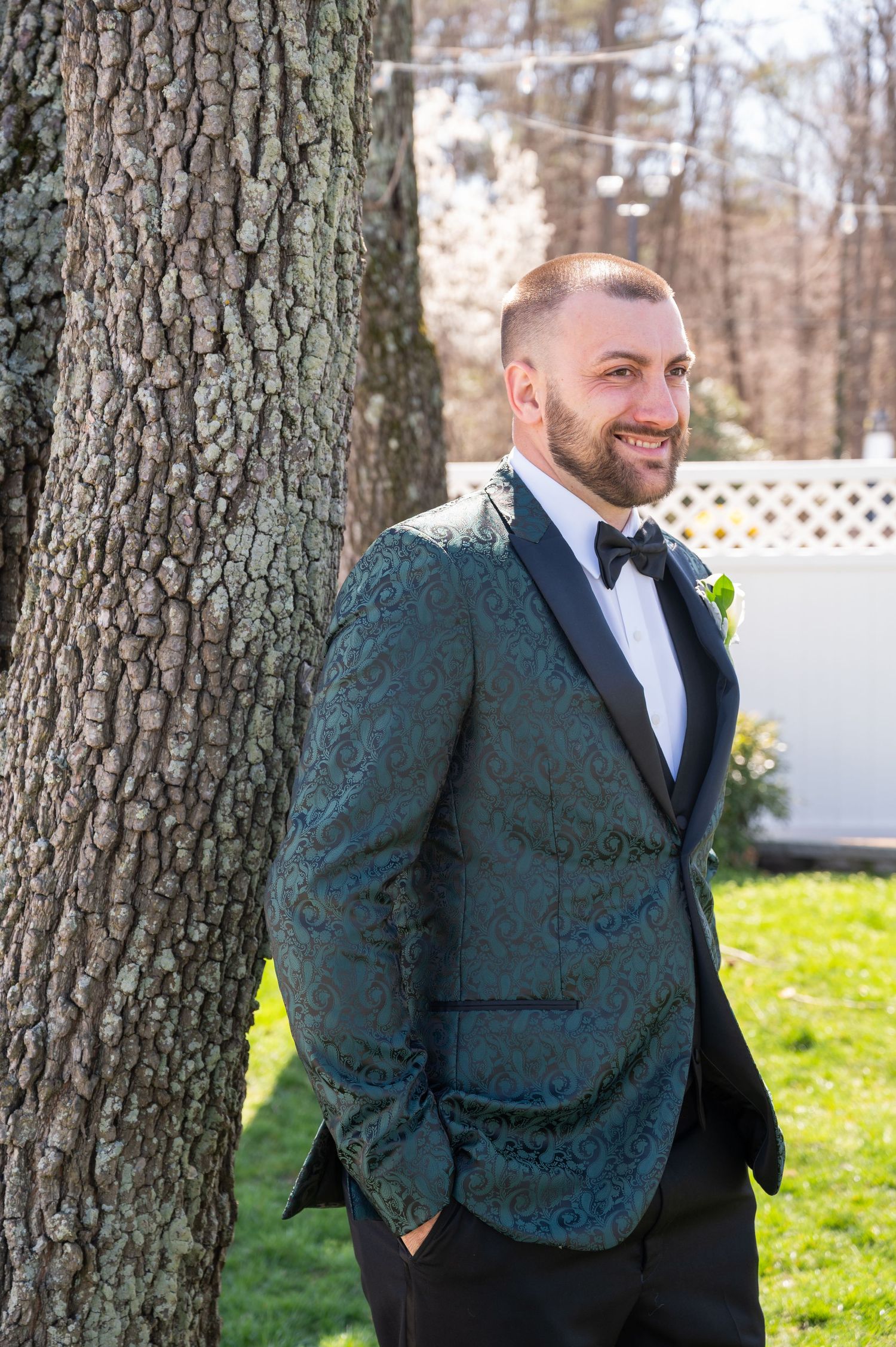 Smiling groom in teal paisley tuxedo jacket leans against tree in outdoor wedding setting.