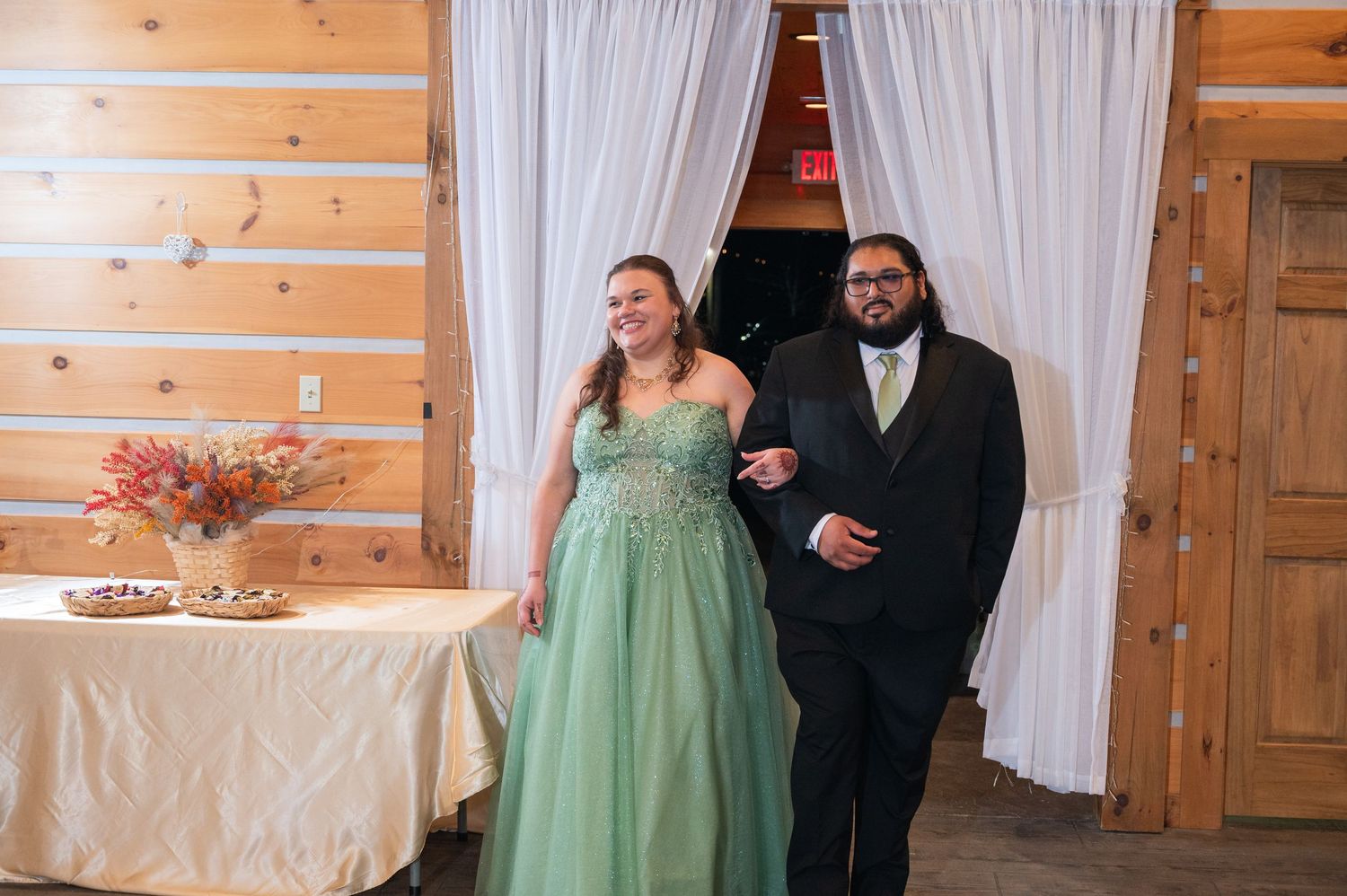 Bride in mint green ball gown and groom in black suit enter rustic barn wedding reception venue together.