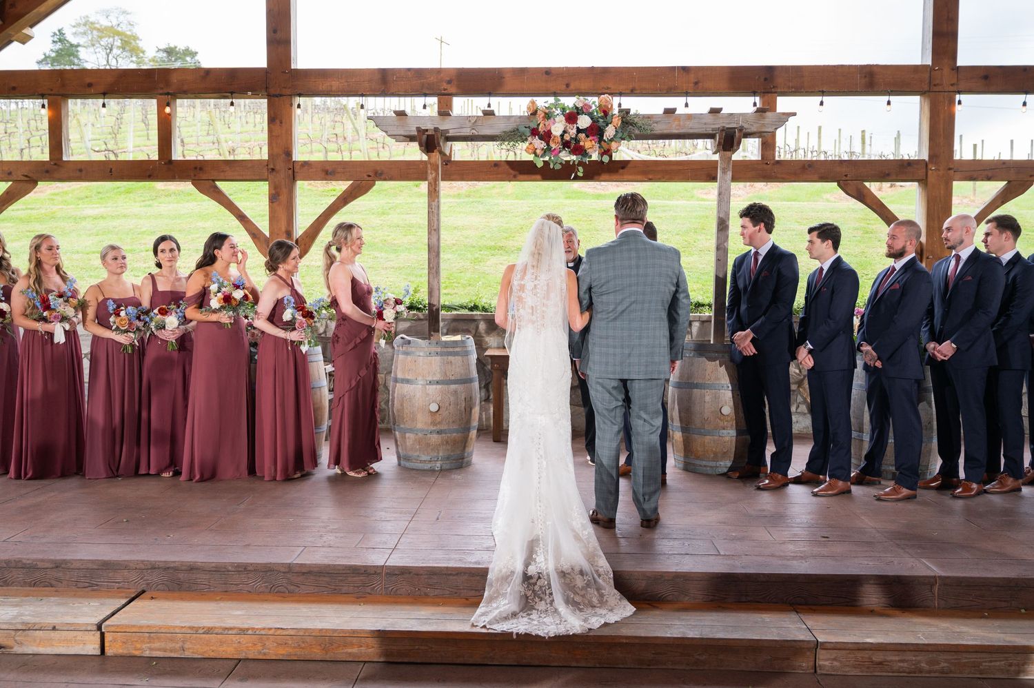Wedding ceremony at rustic barn venue with bridesmaids in burgundy dresses and groomsmen standing at floral altar.