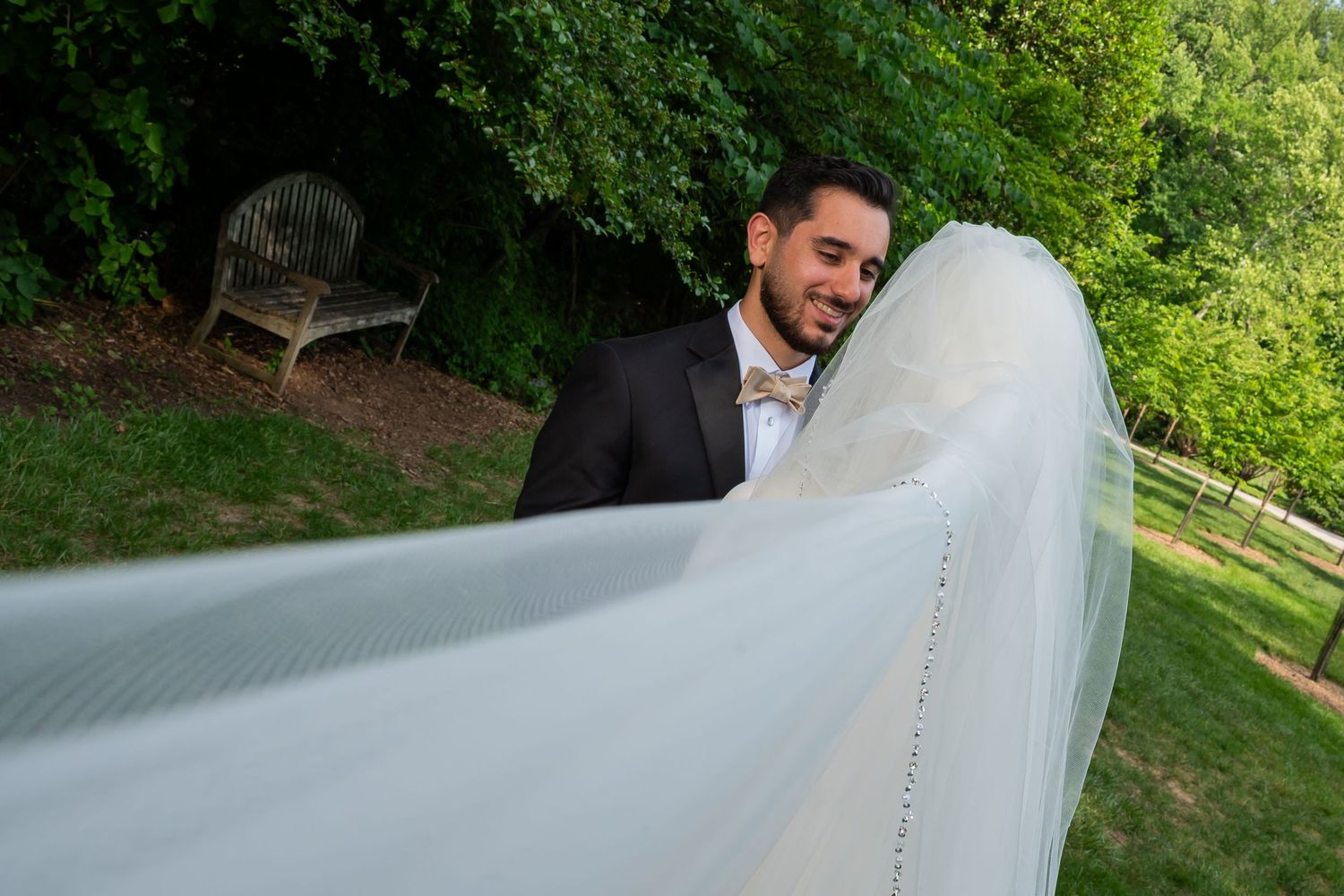 Groom in black suit smiles as bride's white veil billows in the breeze during outdoor wedding ceremony.