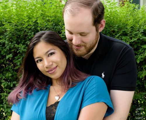 Couple embracing outdoors in front of green hedges, woman with purple-tipped hair smiling.