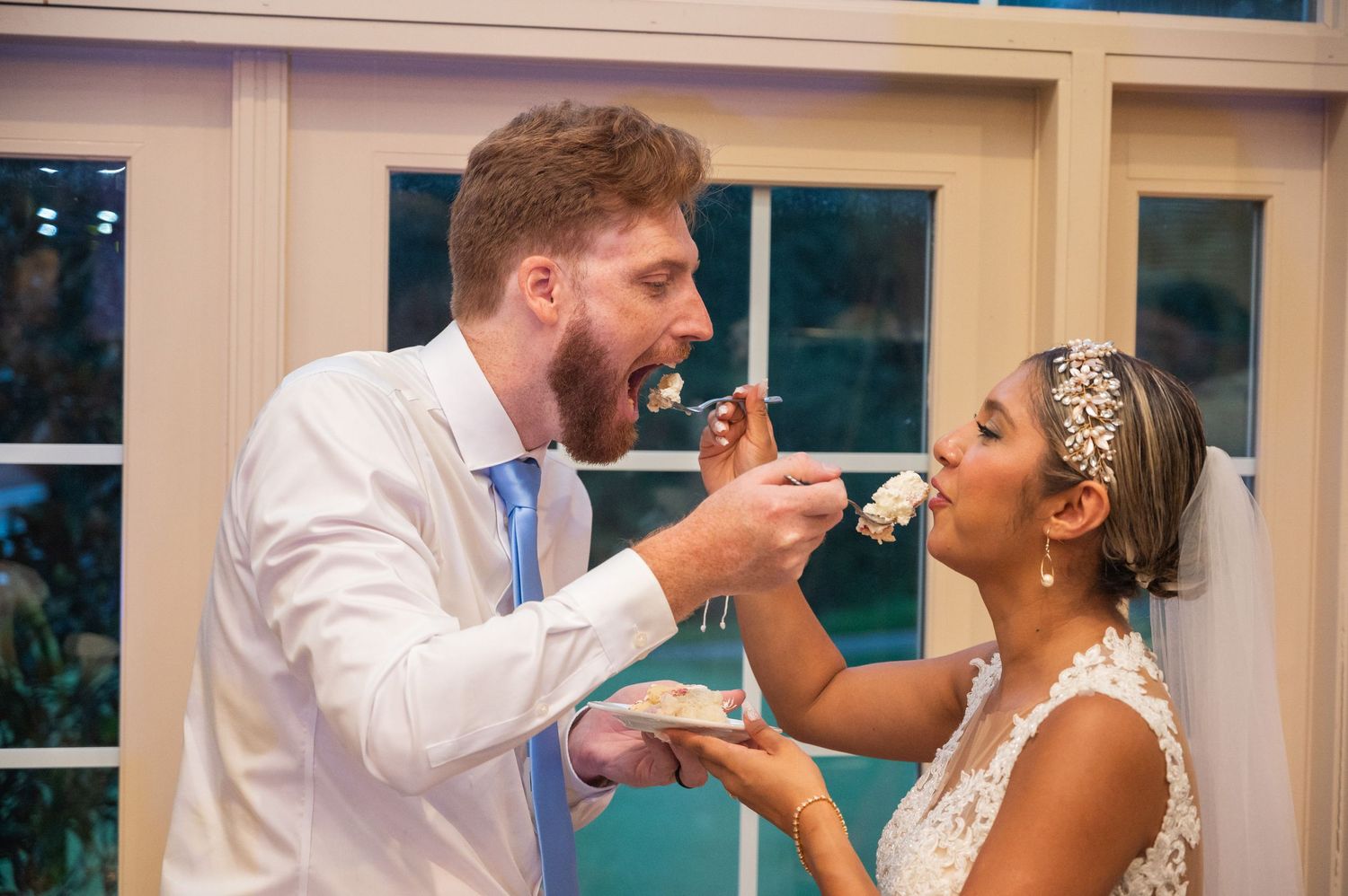 Bride and groom feeding each other wedding cake at their reception, smiling joyfully in elegant attire.
