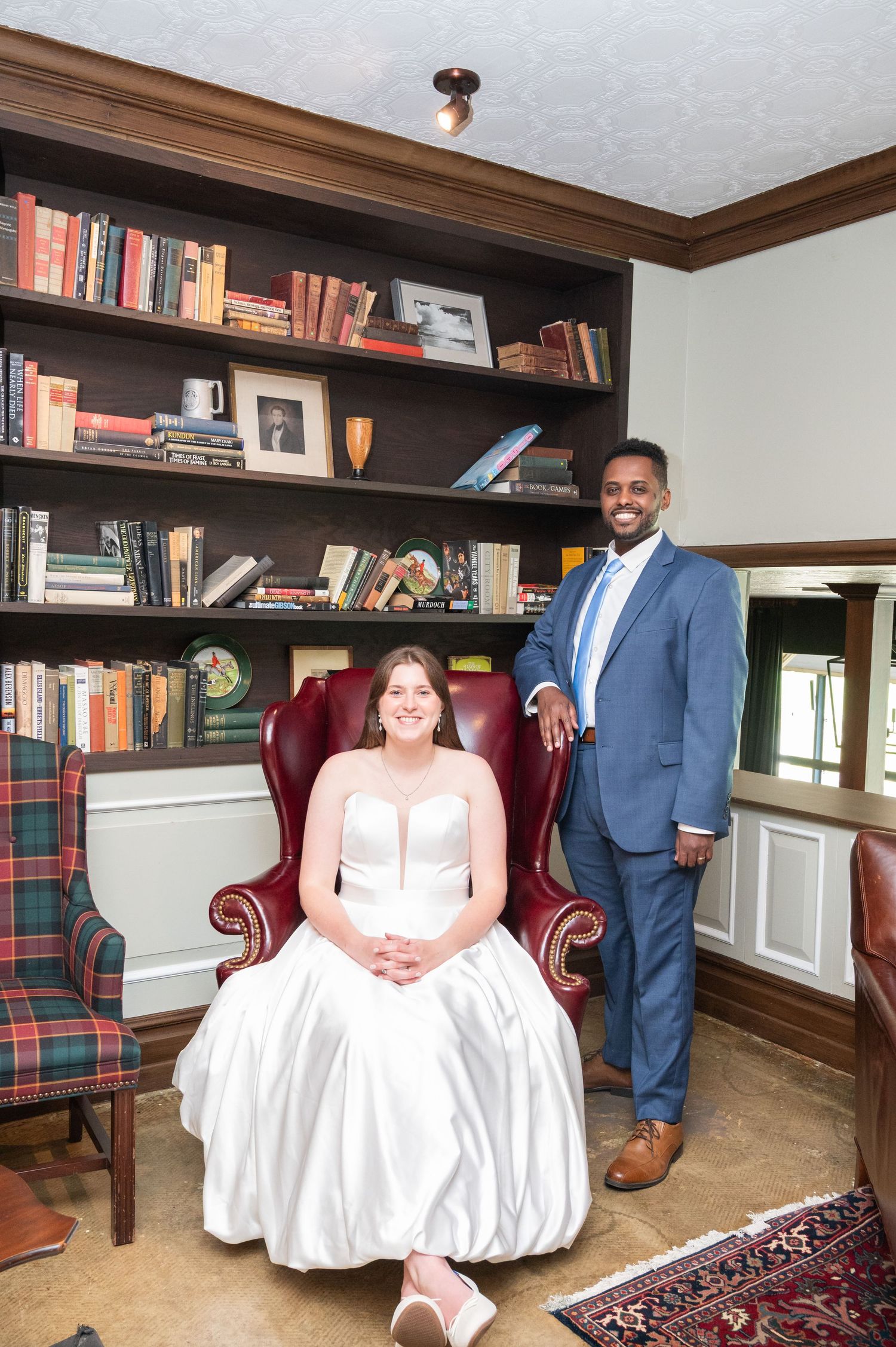 Bride in white gown seated in red leather chair with groom standing beside bookshelf.