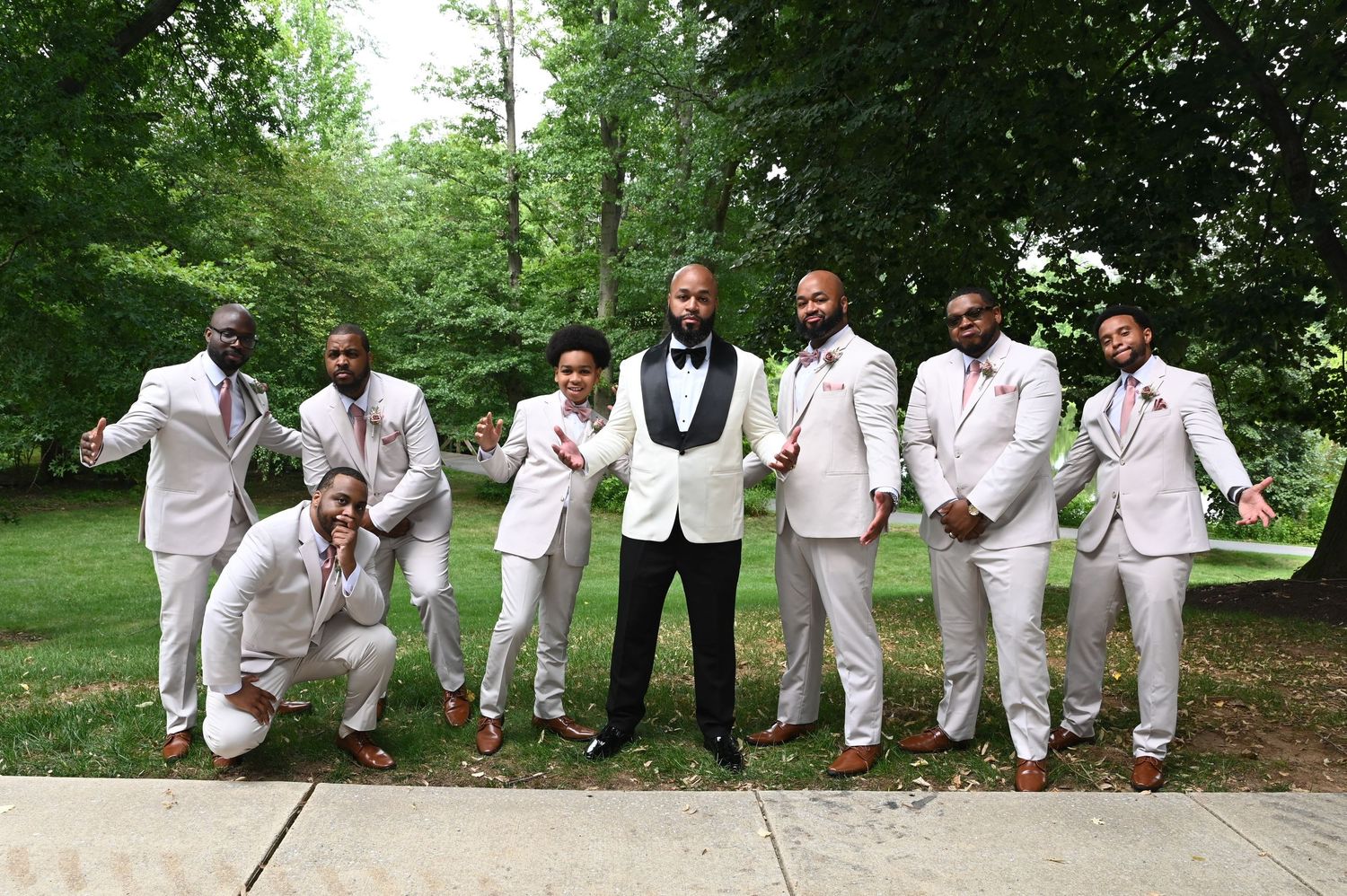 Groom in black tuxedo poses with seven groomsmen in white suits outdoors at a wedding celebration.