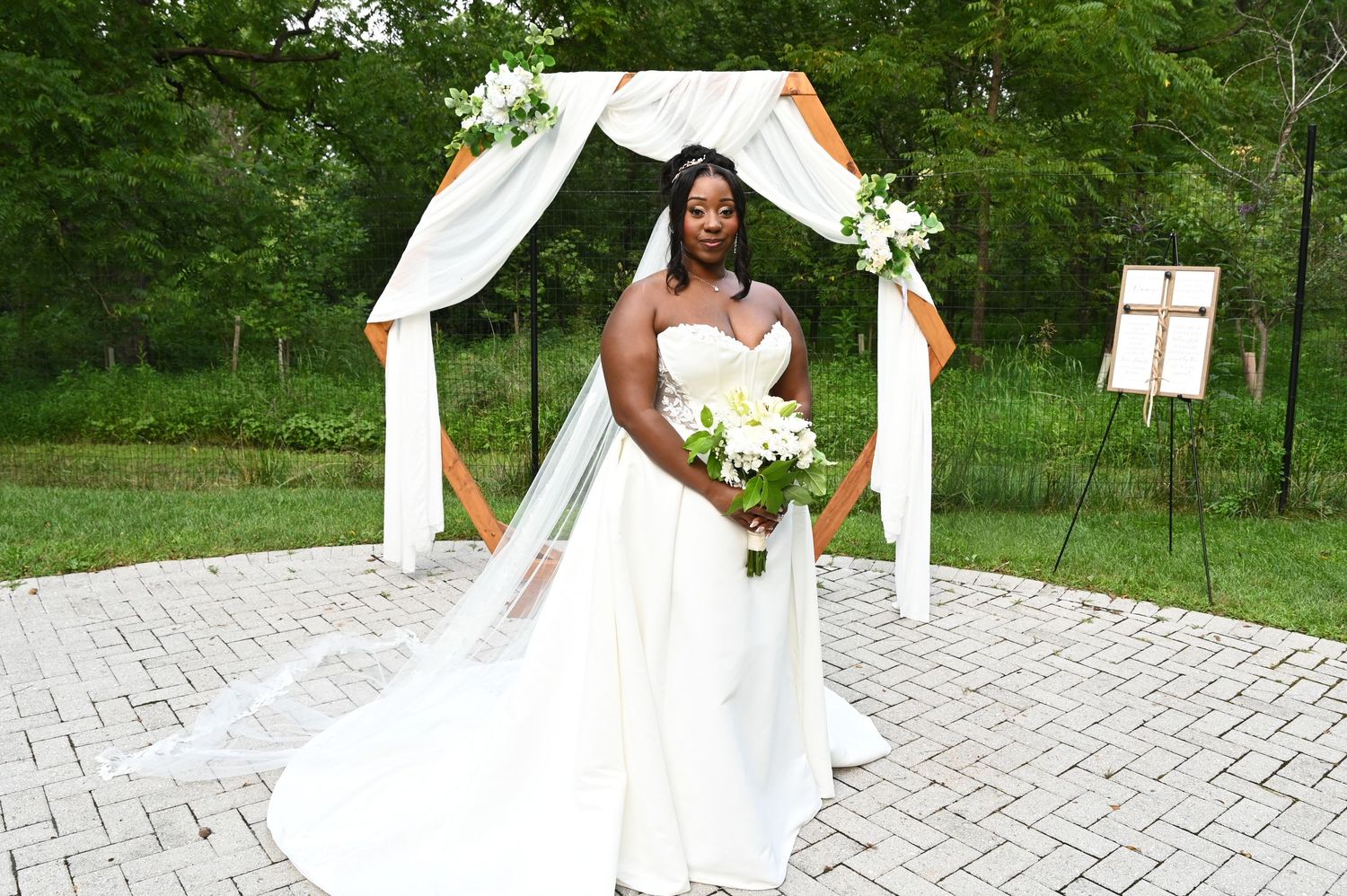 Bride in white strapless gown with veil holds bouquet under floral arch at outdoor wedding ceremony.