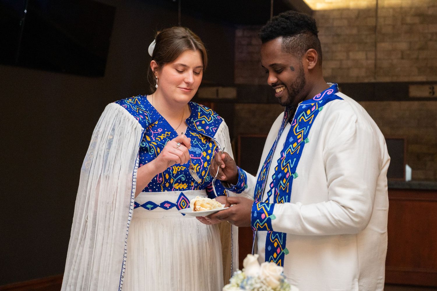 Bride and groom in matching blue and white Ethiopian traditional attire sharing wedding cake together.
