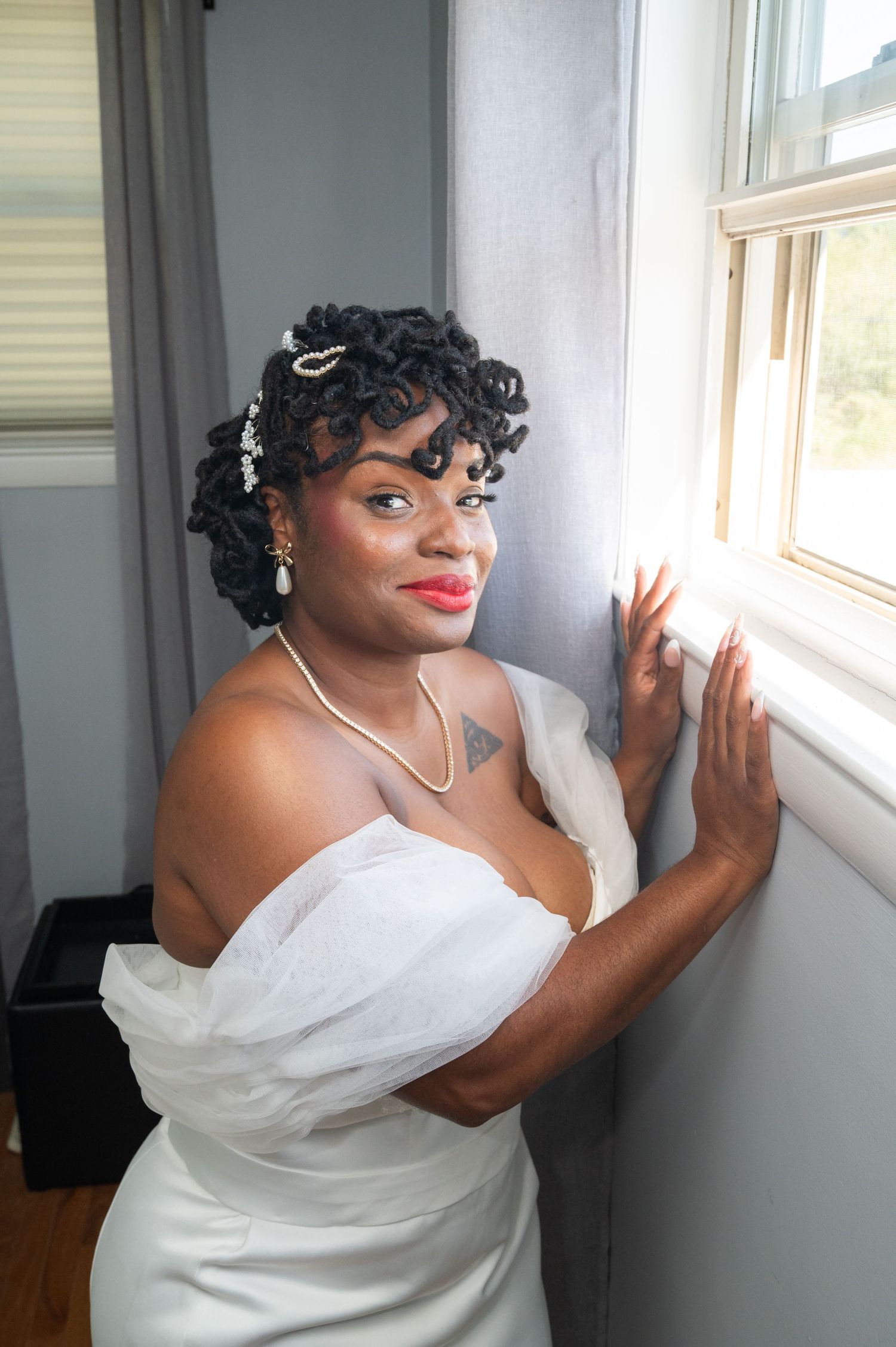 Smiling bride in white off-shoulder gown with curly updo posing by a window indoors.