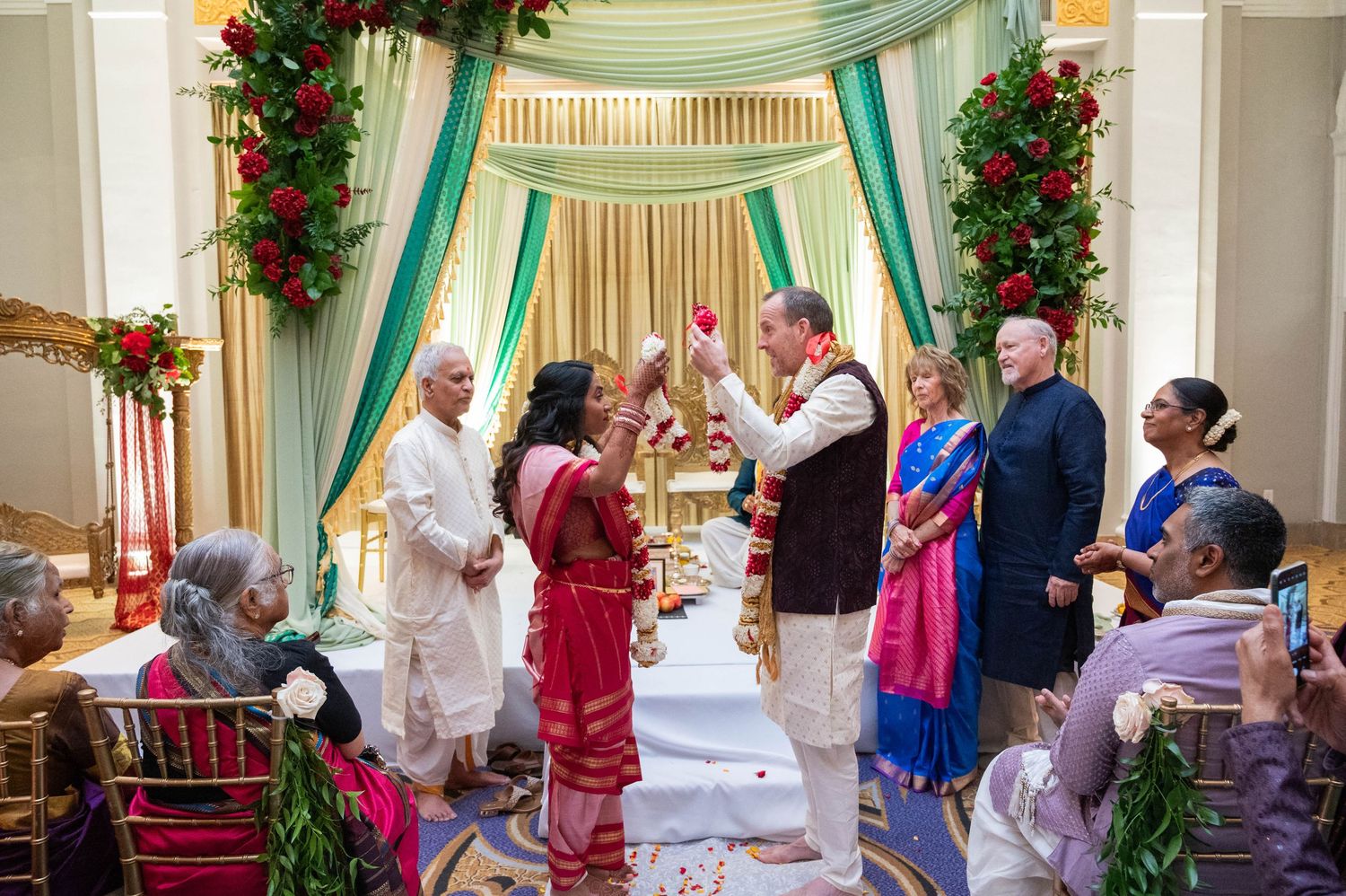 Couple standing under decorated mandap with red flowers during traditional Hindu wedding ceremony indoors.