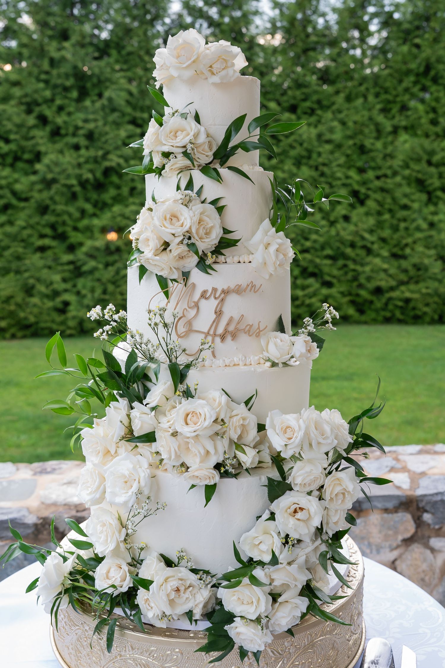 Elegant four-tier white wedding cake adorned with white roses and greenery with gold name topper.