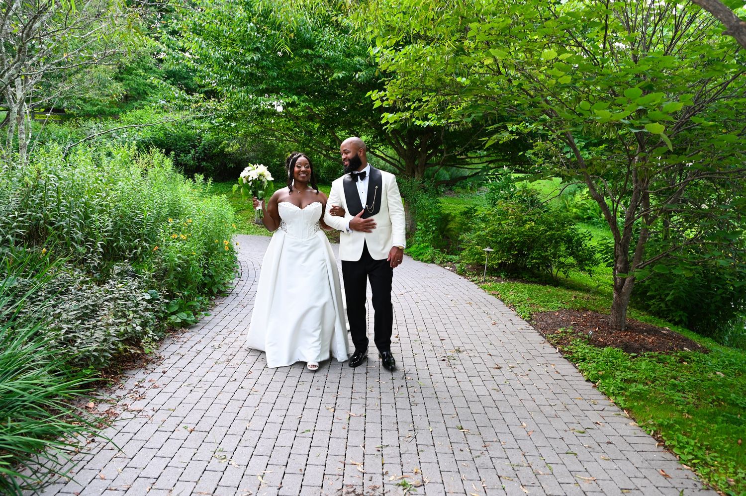 A bride and groom walk hand-in-hand along a brick path surrounded by lush green gardens on their wedding day.