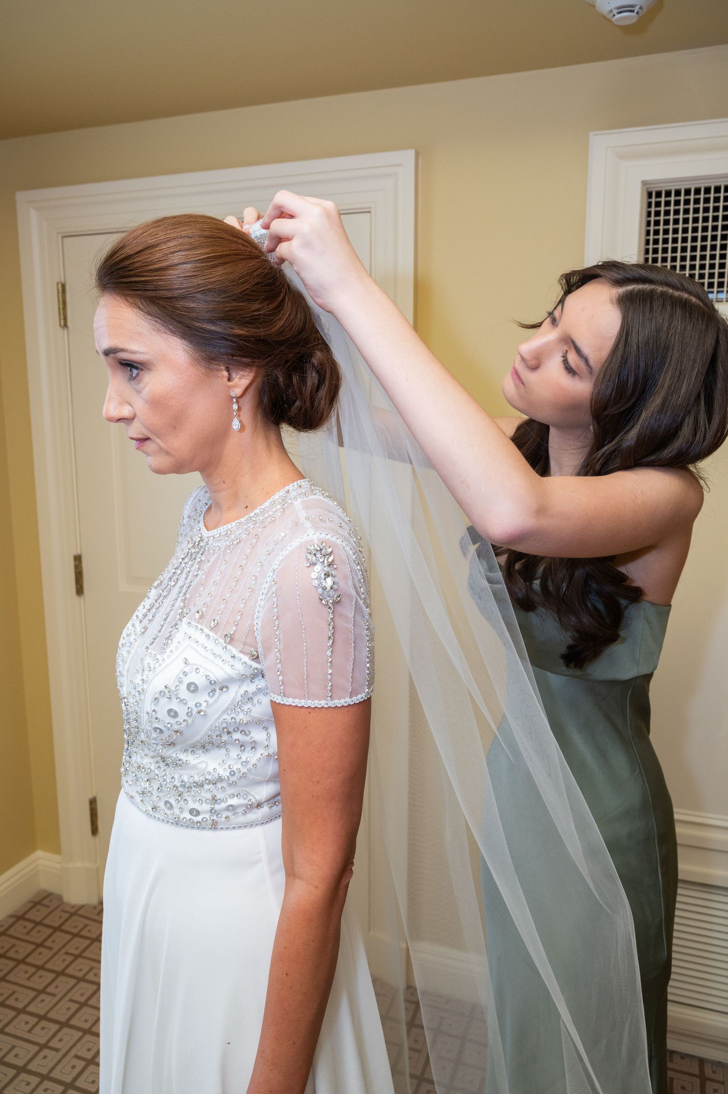 Bridesmaid carefully placing veil into bride's elegant updo in a warmly lit room.