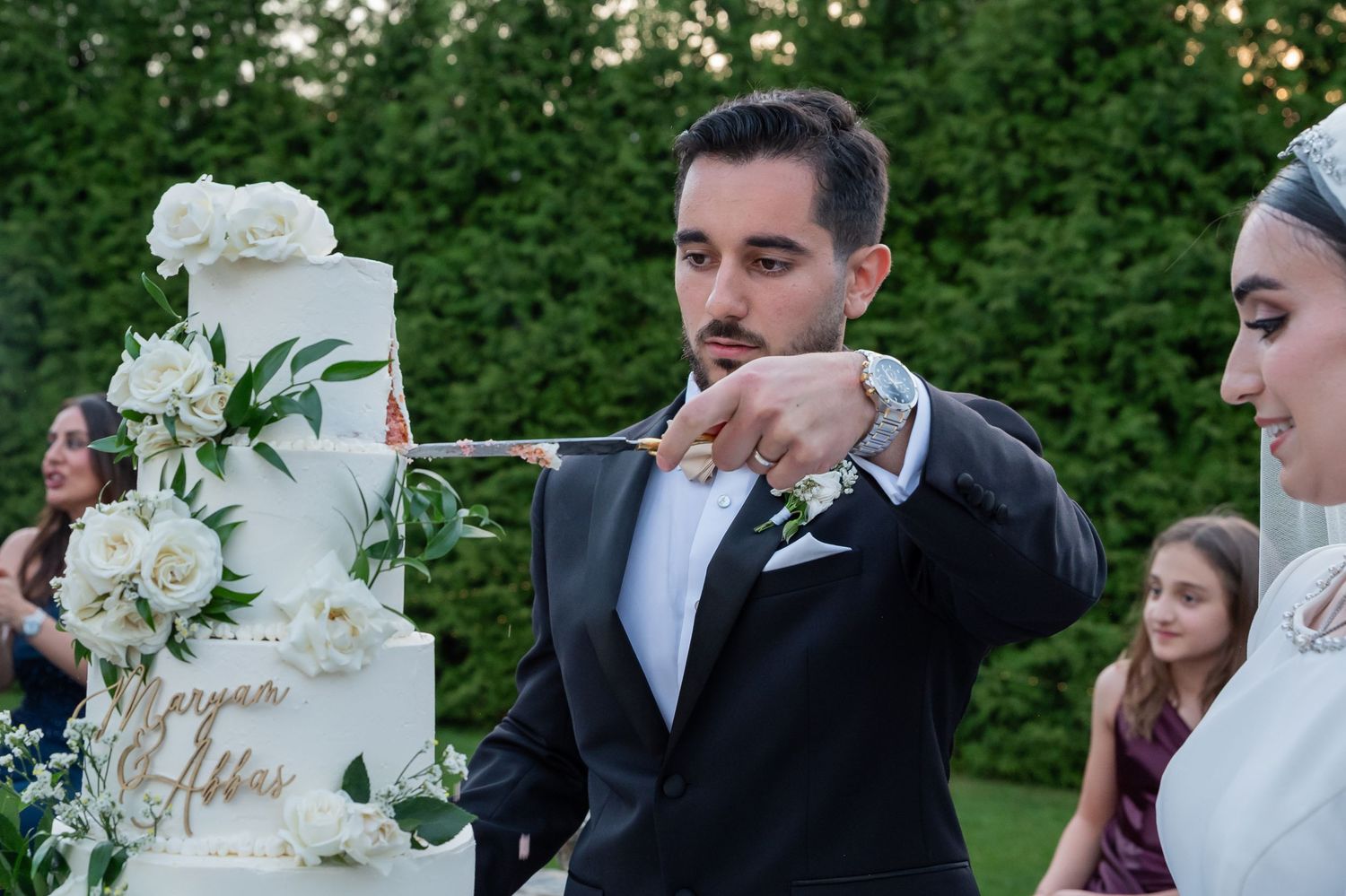 Bride and groom cutting a white floral wedding cake outdoors at an elegant garden wedding ceremony.