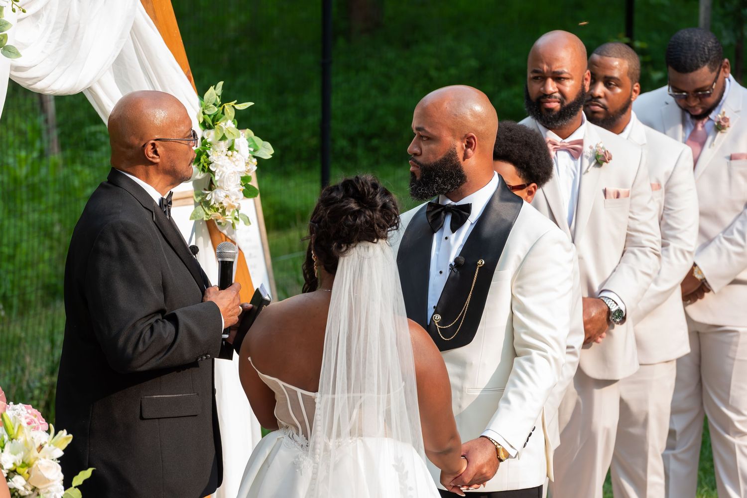 Groom in white tuxedo exchanges vows with bride in wedding gown, flanked by groomsmen in blush suits outdoors.