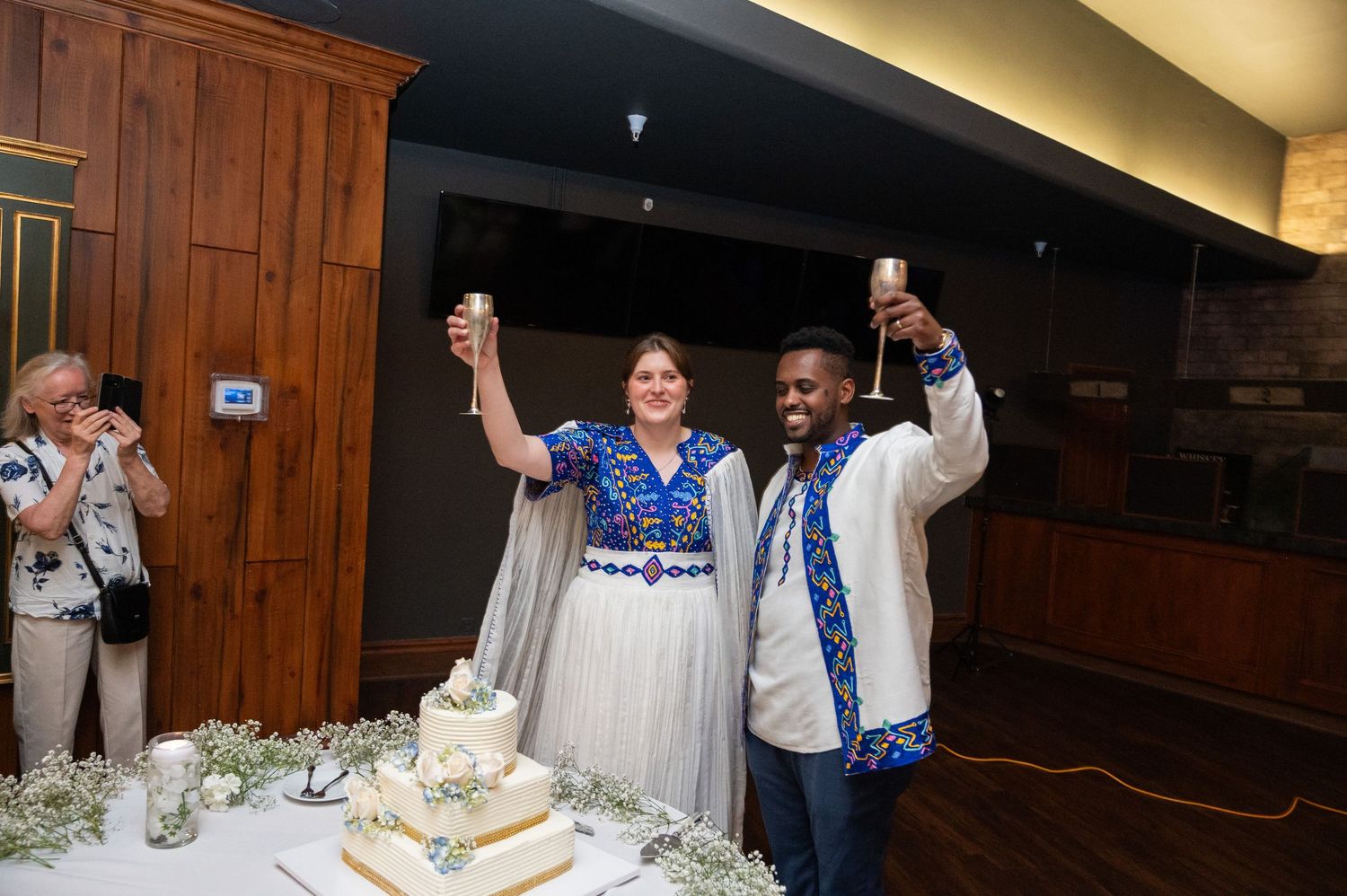 Bride and groom in traditional Ethiopian wedding attire toasting with champagne glasses beside wedding cake.
