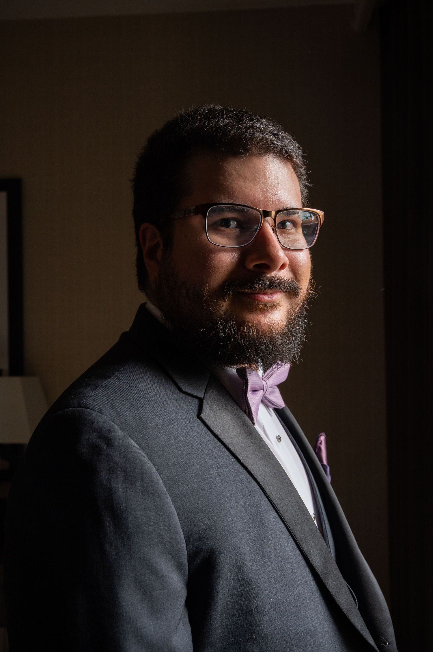 Dapper groom in gray suit with lavender bow tie and glasses posing in dramatic lighting.