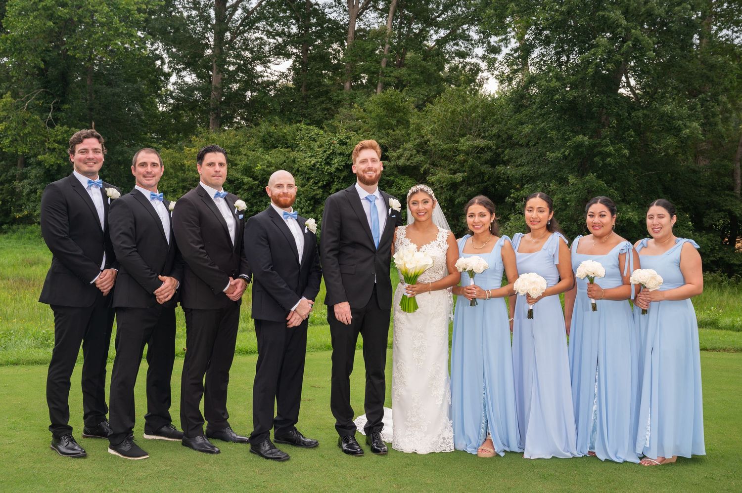 Wedding party poses on golf course with groomsmen in black tuxedos and bridesmaids in light blue gowns.