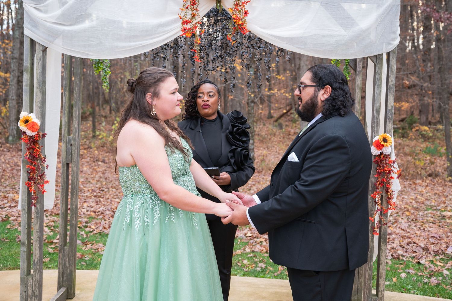 Couple exchanges vows in outdoor woodland wedding ceremony with officiant, bride in green gown, groom in black suit.