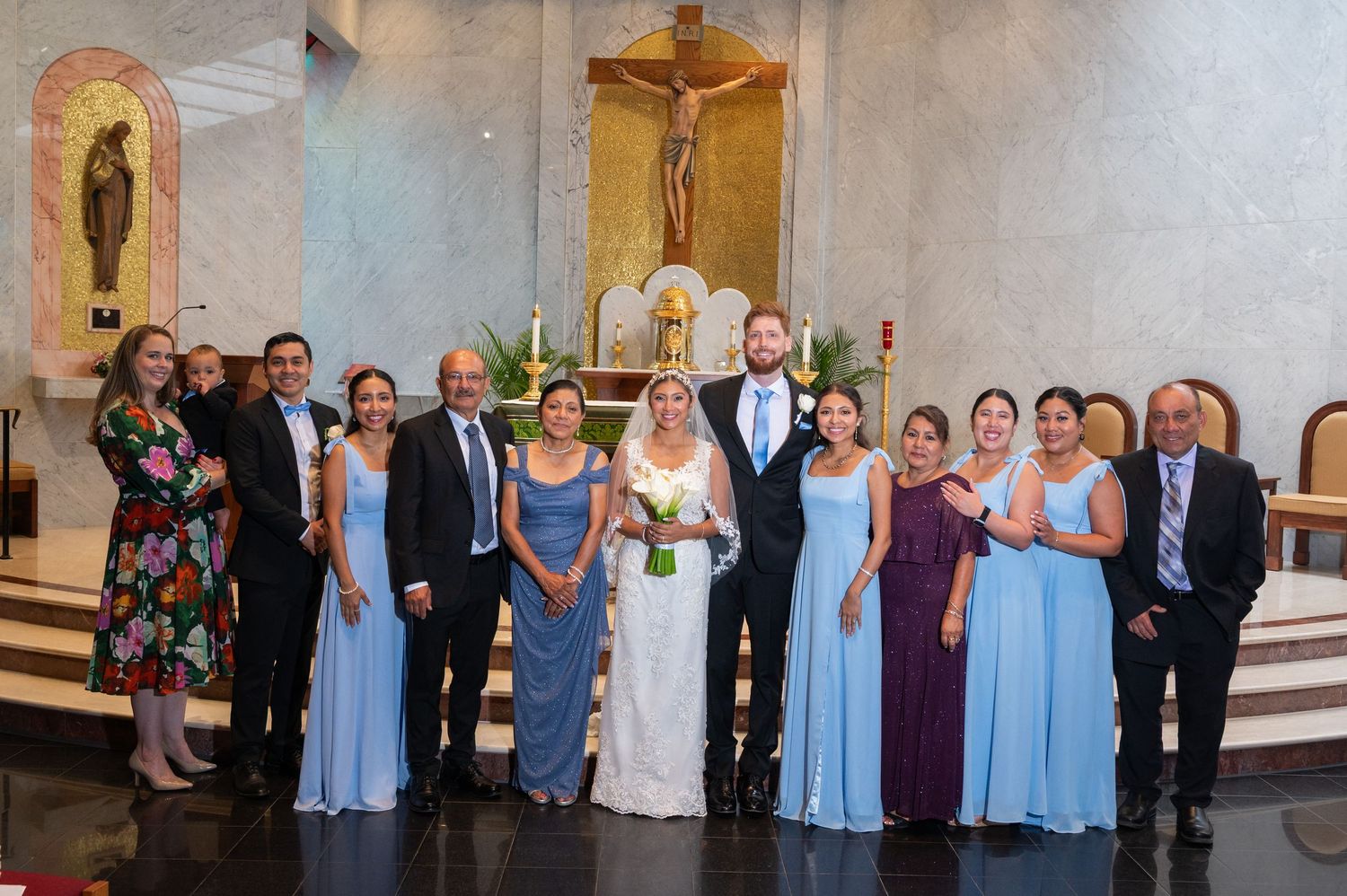 Wedding party poses in church with bride and groom, bridesmaids in blue dresses, before altar with crucifix.